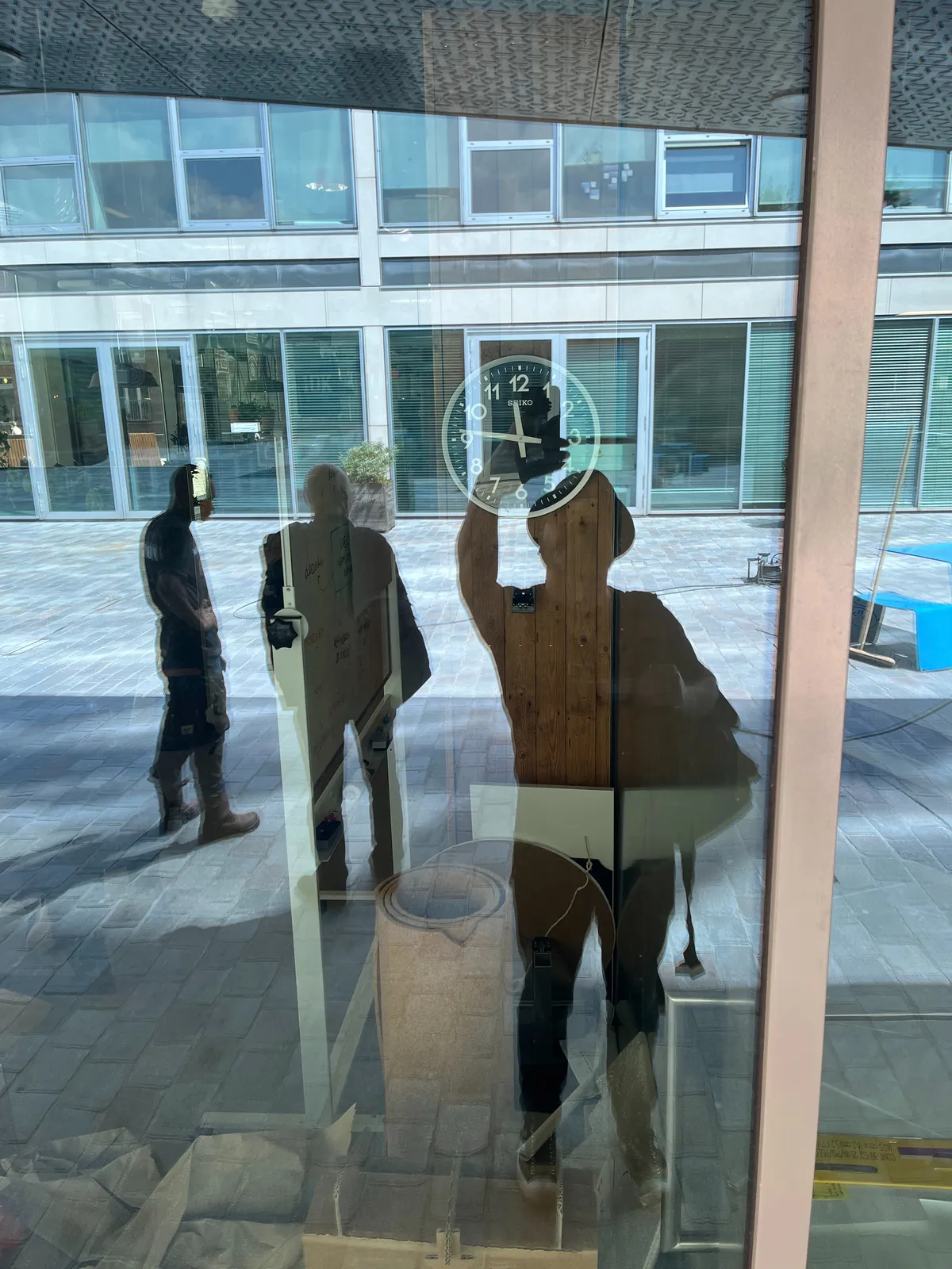 In the reflection of a large window three people can be seen. One of them takes this picture. Through the window a clock can be seen which points at 14 minutes to 12.