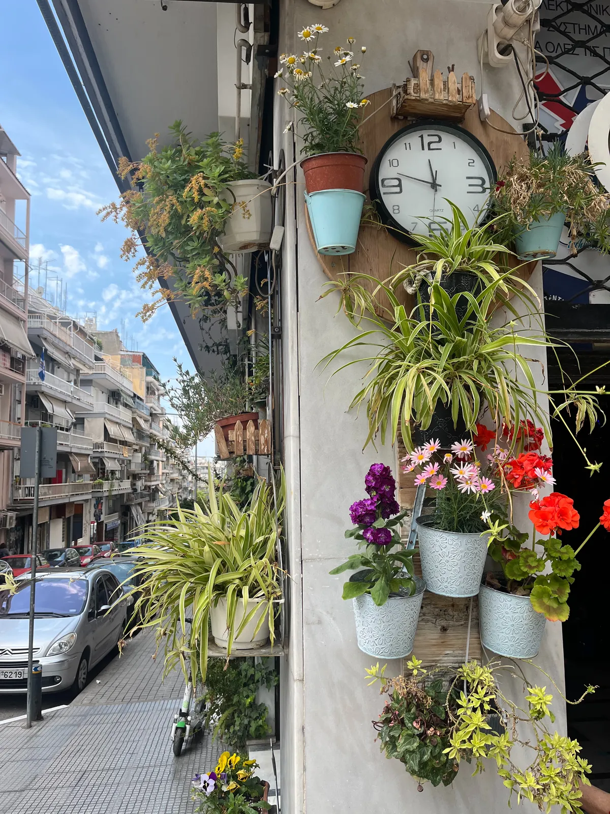 Looking down a street with Greek appartment buildings, past the corner of a shop. There are many flowers, and a clock on this shop’s facade. It looks nice. The clock points at 13 minutes to twelve.