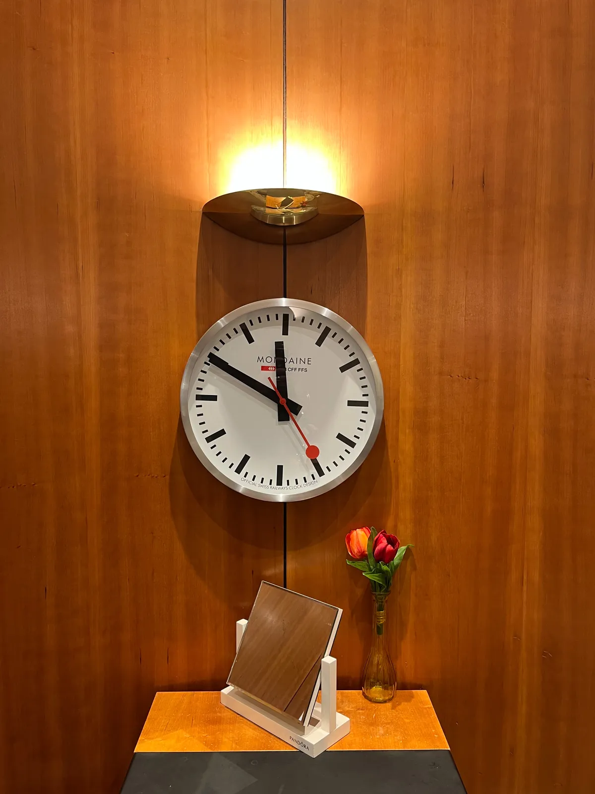 A classic Swiss wall clock, with two black hands, and a bright red second hand. It is attached to a wooden wall, below a silver wall light, and above a square mirror, and a vase with a rose in it. The clock points at 11 minutes to twelve. 