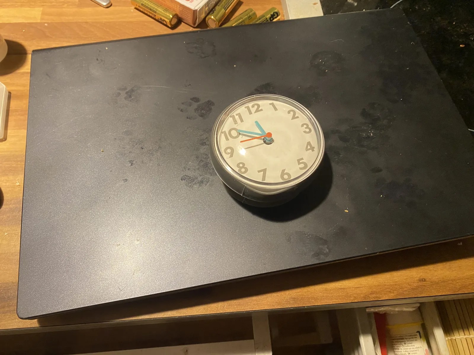 A clock on a dusty black plank. You can see that a cat walked over it. The clock points at 9 minutes to 12.