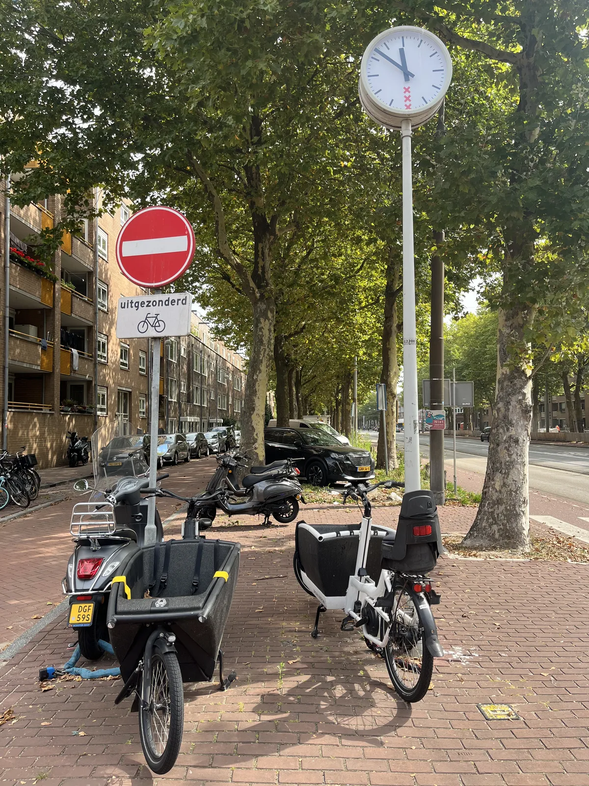 Two cargo carrier bikes and some scooters parked on a street, lined with green trees. In front of the trees there’s a sign that says no entry, except for bycicles, and there’s a clock on a pole that points at 8 minutes to twelve.