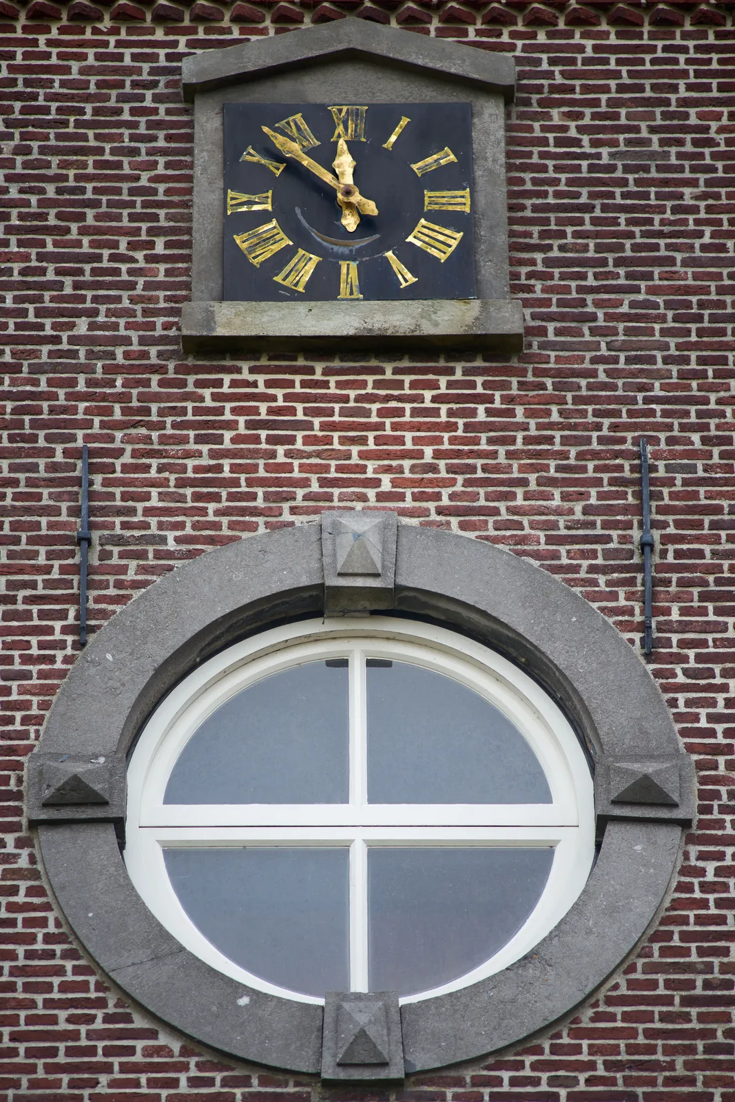 A clock on a brick wall with a round window below it. The clock points at 7 minutes to 12.