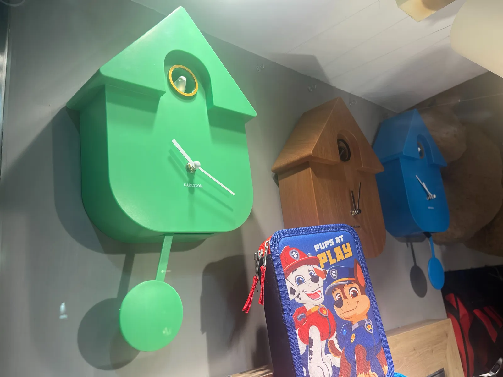 Three modern cuckoo clocks. The brown one in the middle points at four minutes past twelve. The bright green one on the left points at approximately 19 minutes past 10 and the blue one on the right points at 6 minutes to twelve.