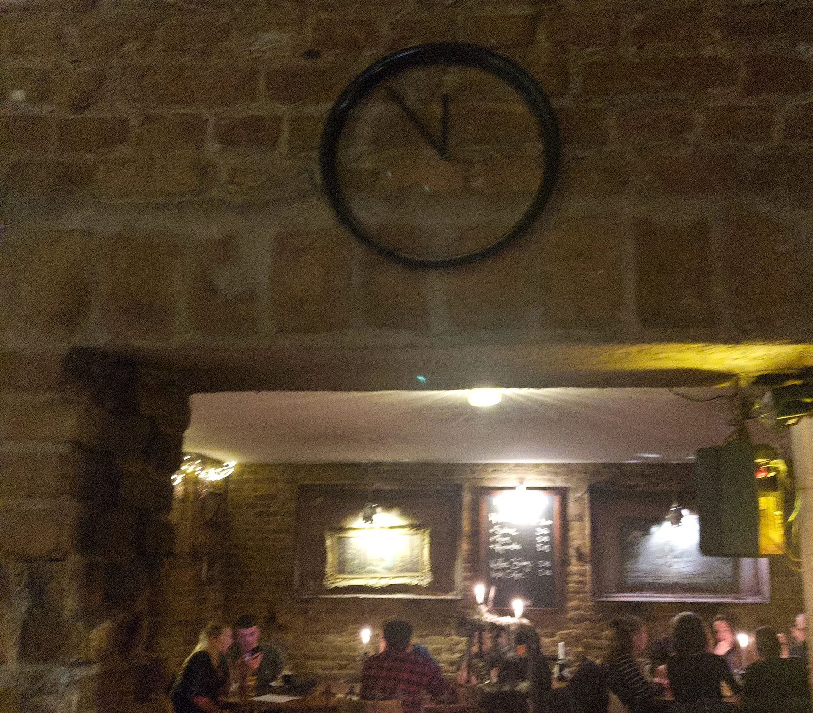 Peeking through a door opening, in the background there are people sitting at tables. Above the door, on a brick wall, there’s a minimalist clock that points at approximately 6 minutes to 12.