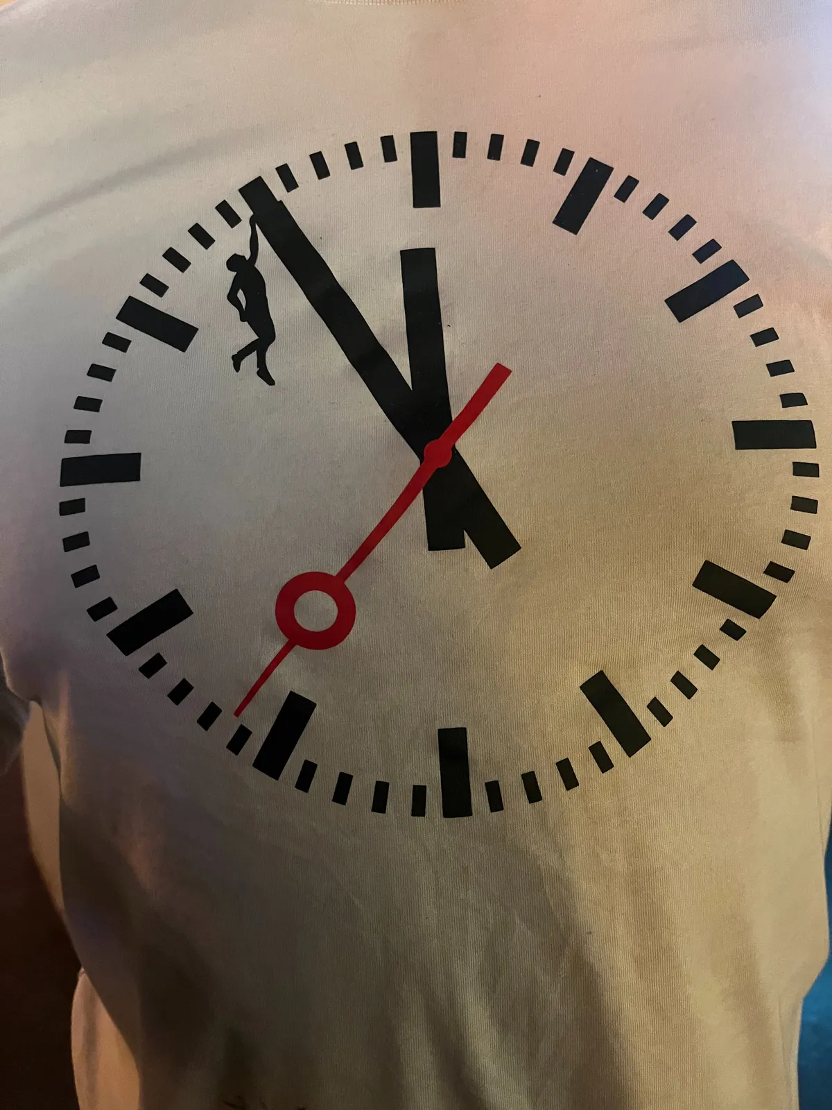 A clock printed on a t-shirt. A person is hanging, holding onto the minute hand. The clock points at five minutes to 12.