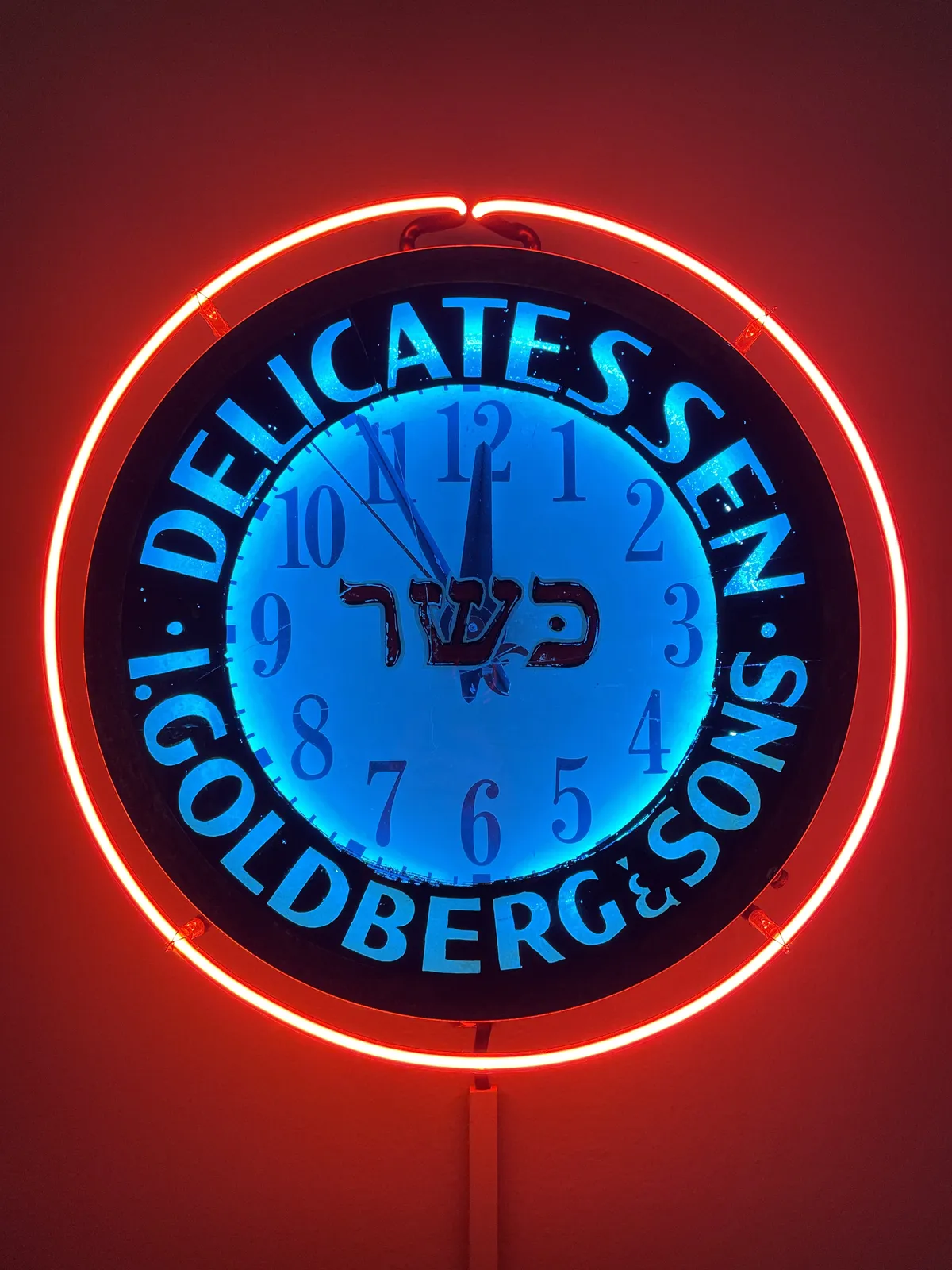 A colourful, neon lit clock, that says Delicatessen I Goldberg and Sons. It points at 5 minutes to 12.