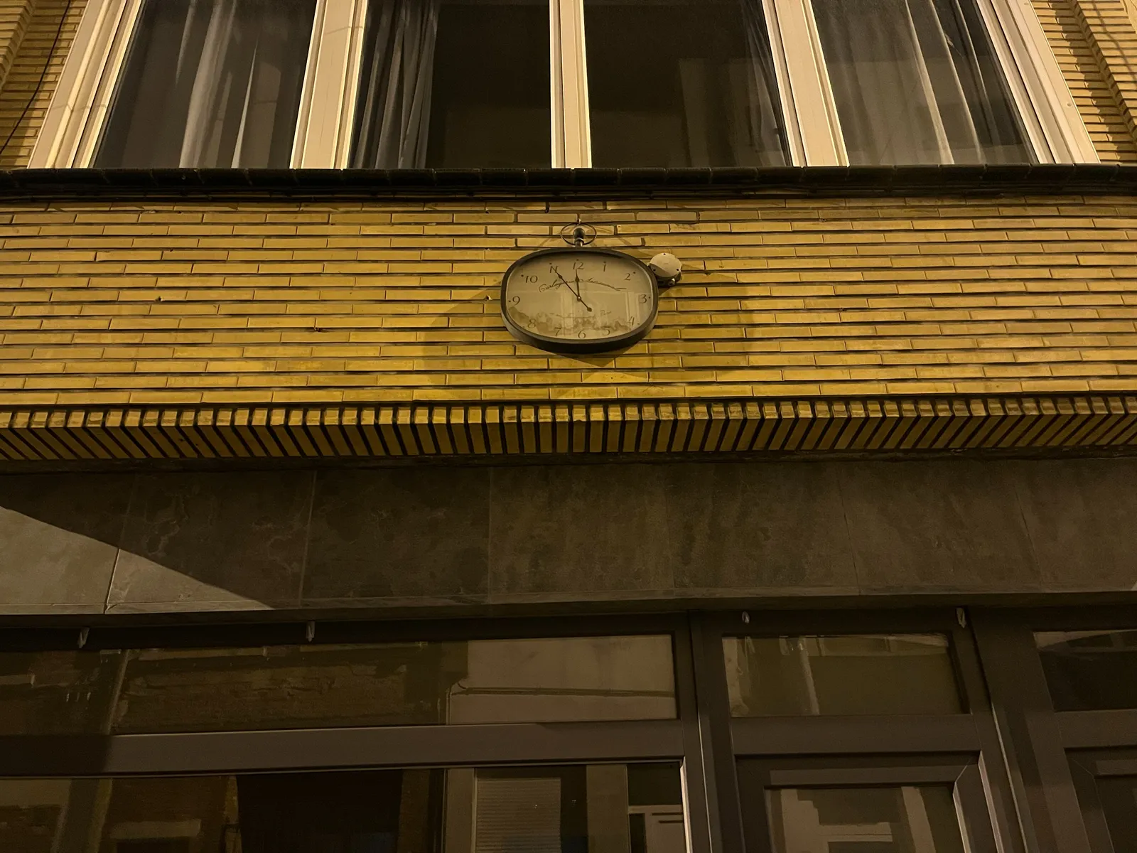 A rather fragile looking clock on a brick wall. It points at 5 minutes to 12.