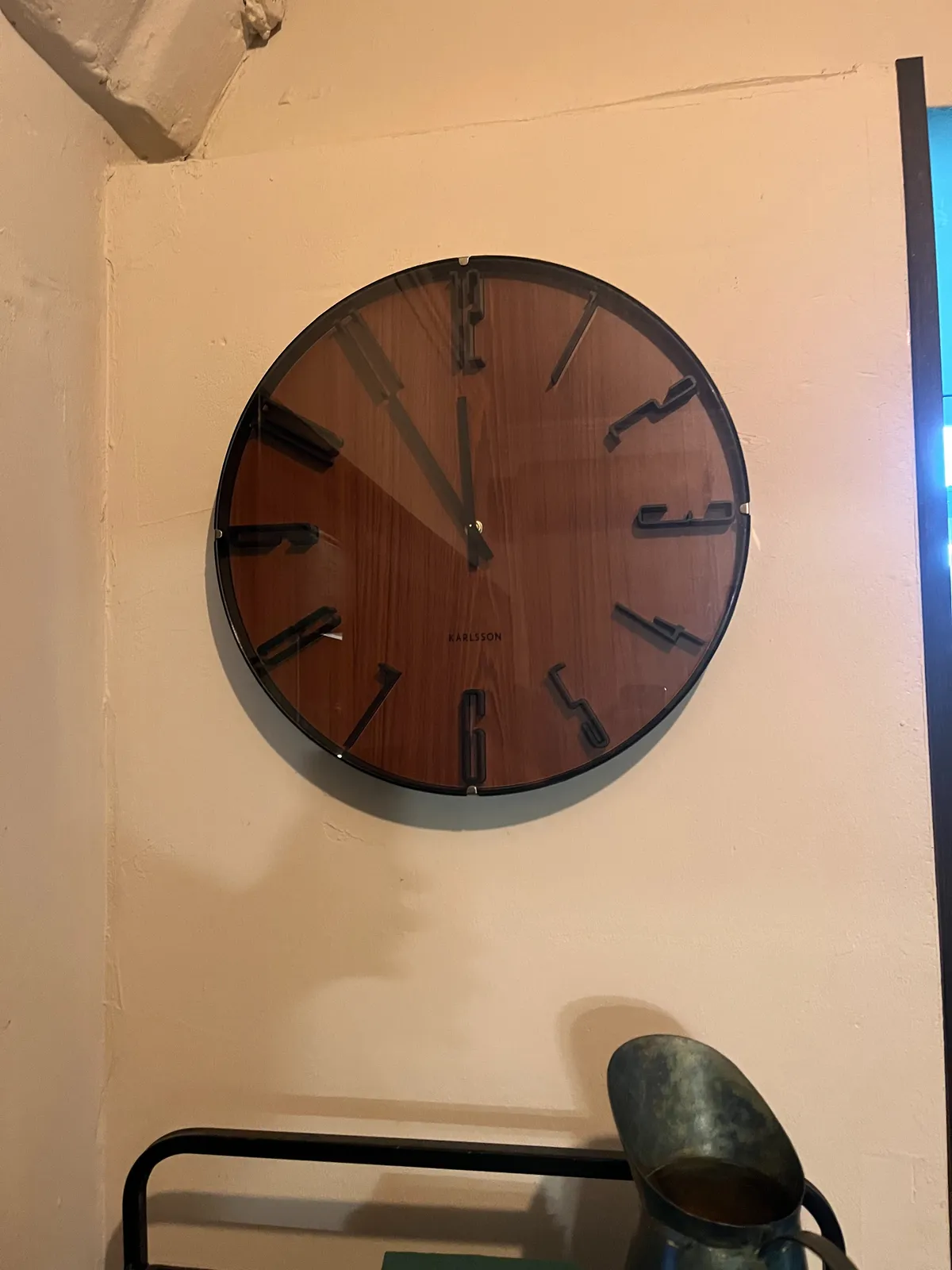 A dark wooden clock, with black numbers and black hands, on a white wall. The clock points at 5 minutes to 12.