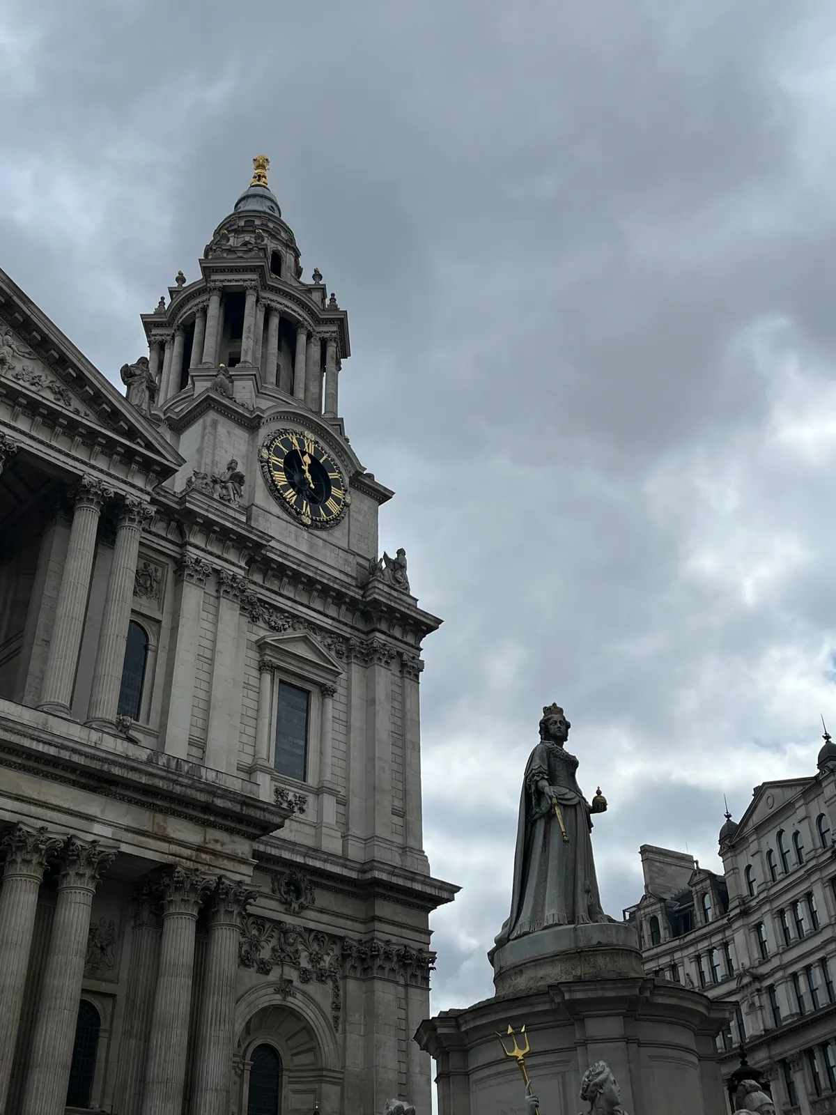A statue of a queen-like figure with behind it an old building with a clock on its tower. It points at 4 minutes to 12.