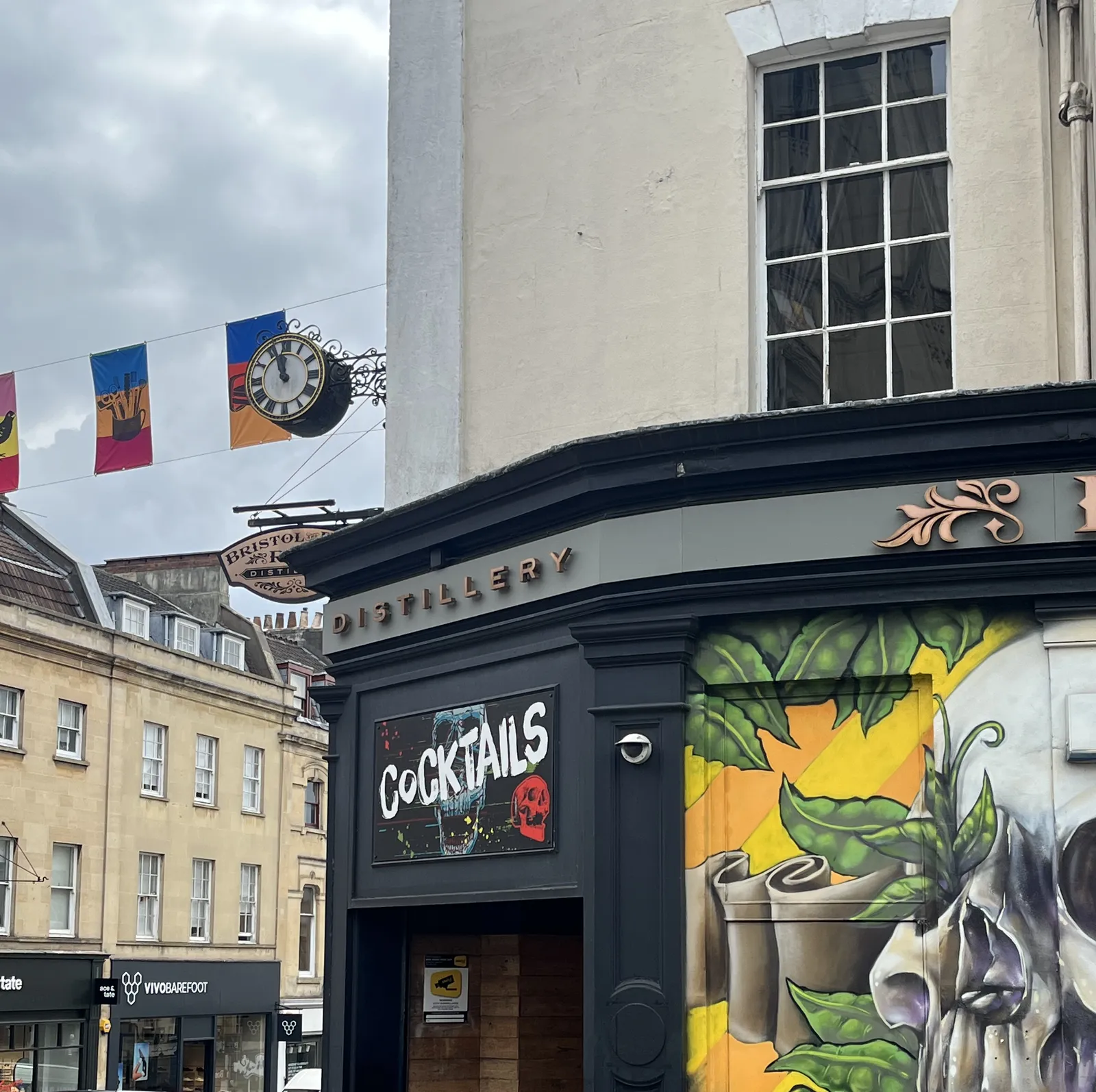 A bar on the corner of the street in, probably, Bristol, since it says so on a sign attached to the bar. There’s a skull painted on the bar, and a clock attached to its facade. It points at three minutes to twelve.