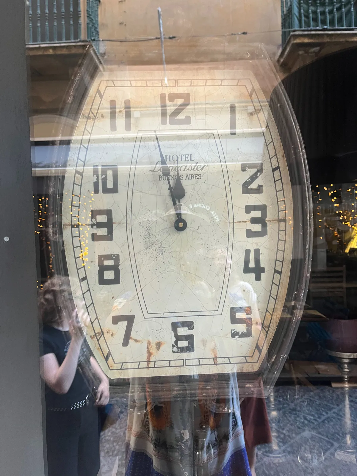 A large clock behind a window. Some balconies and some people can be seen in the reflection. The large clock points at 3 minutes to 12.