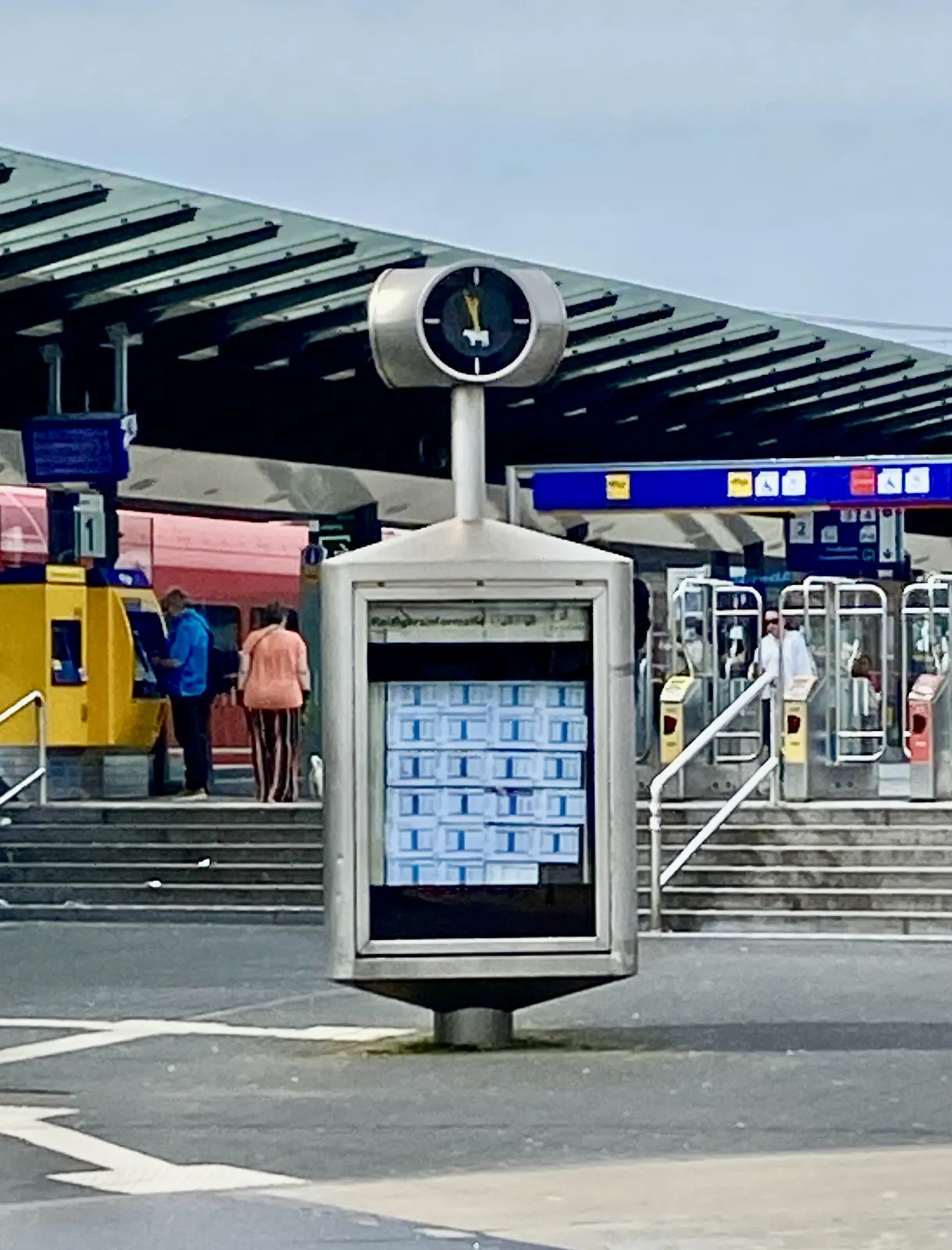 A clock on a square in front of a train station. It points at 2 minutes to 12.