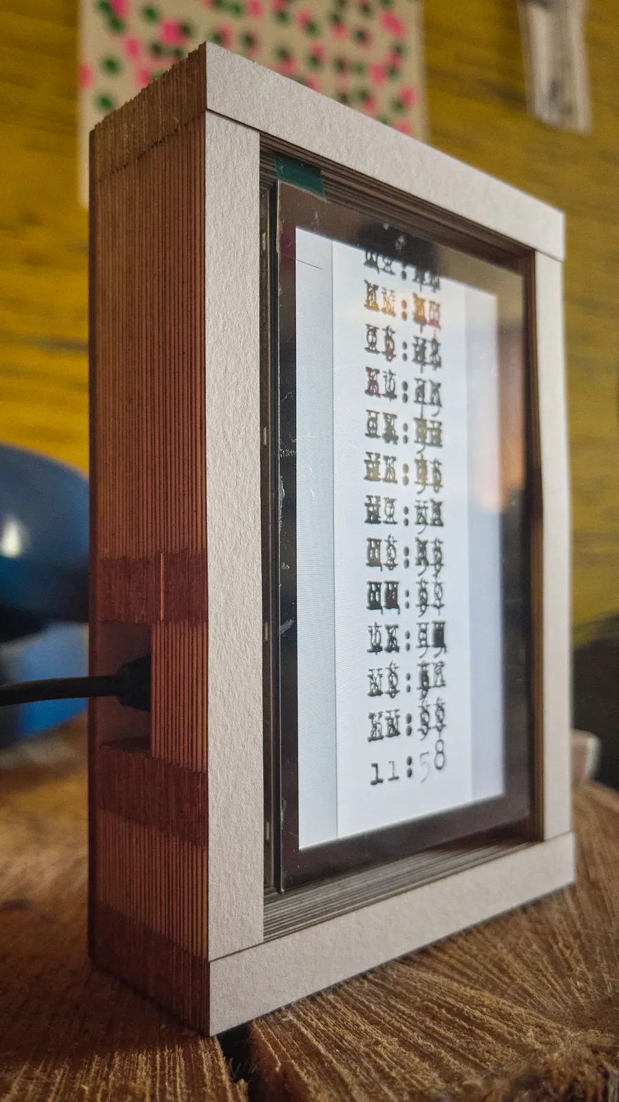 A digital display in a frame made fom many thin layers of paper. On the display there is a list of what seem to be chinese characters, but on closer inspection they turn our to be many types characters over each other. The bottom item in the list is a timestamp which says it is 11:58