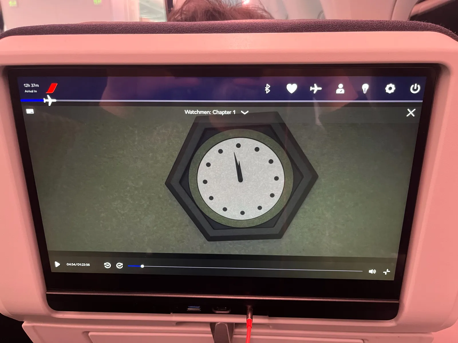 A picture of an in flight entertainment screen, built into a chair. The top of the head of the person sitting in this chair can be seen. The flight is going to take another 12 hours and 37 minutes. The still on the screen is from the movie Watchmen: Chapter one. There’s a close up of a wall clock, which points at 2 minutes to 12.  