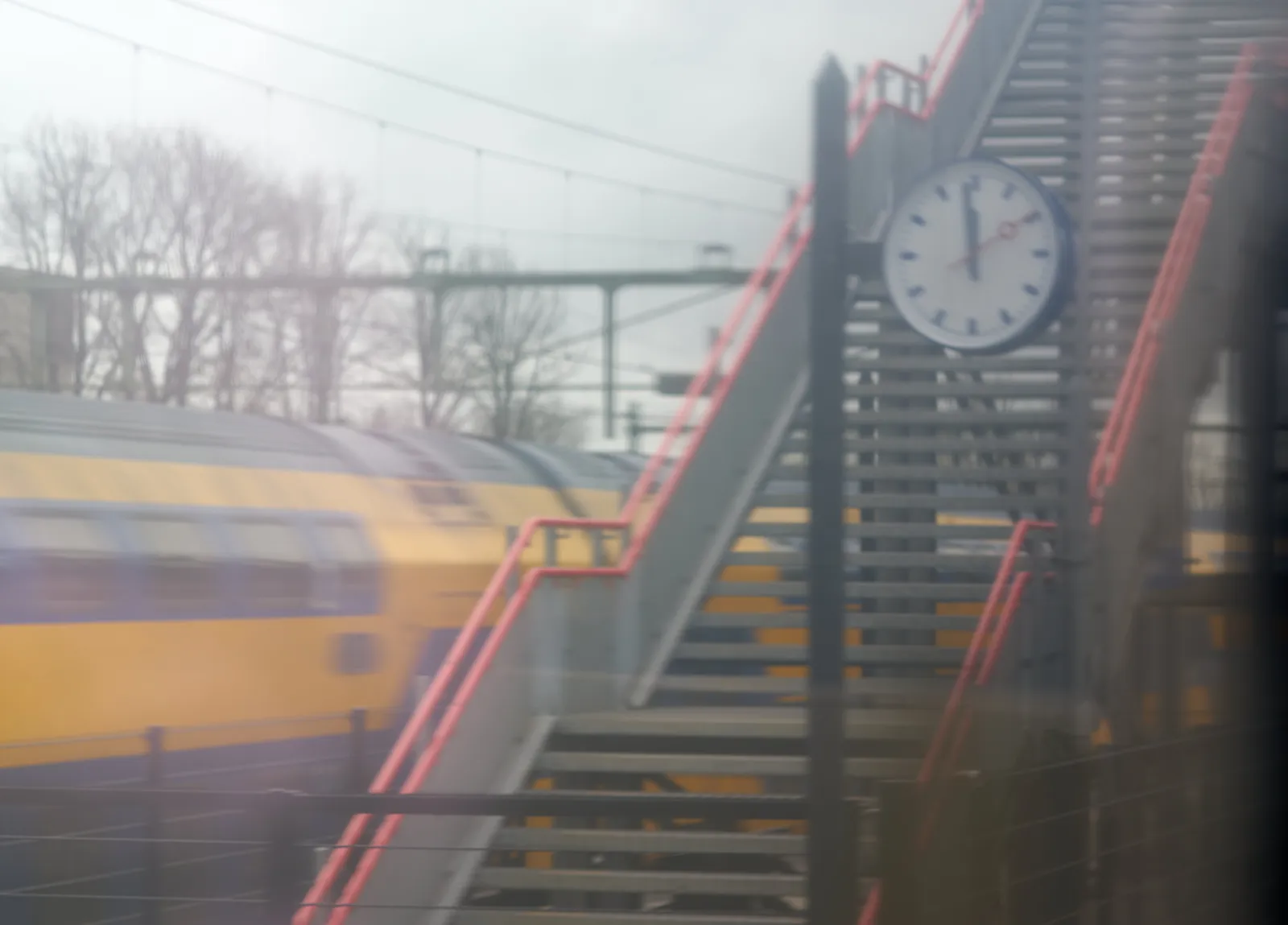 A clock on a train station, next to some stairs. On it it is one minute to 12.
