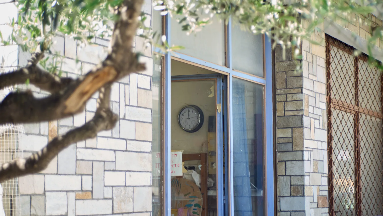 Peeking past a tree, into a workshop, at the clock on the wall. It points at a minute to twelve.
