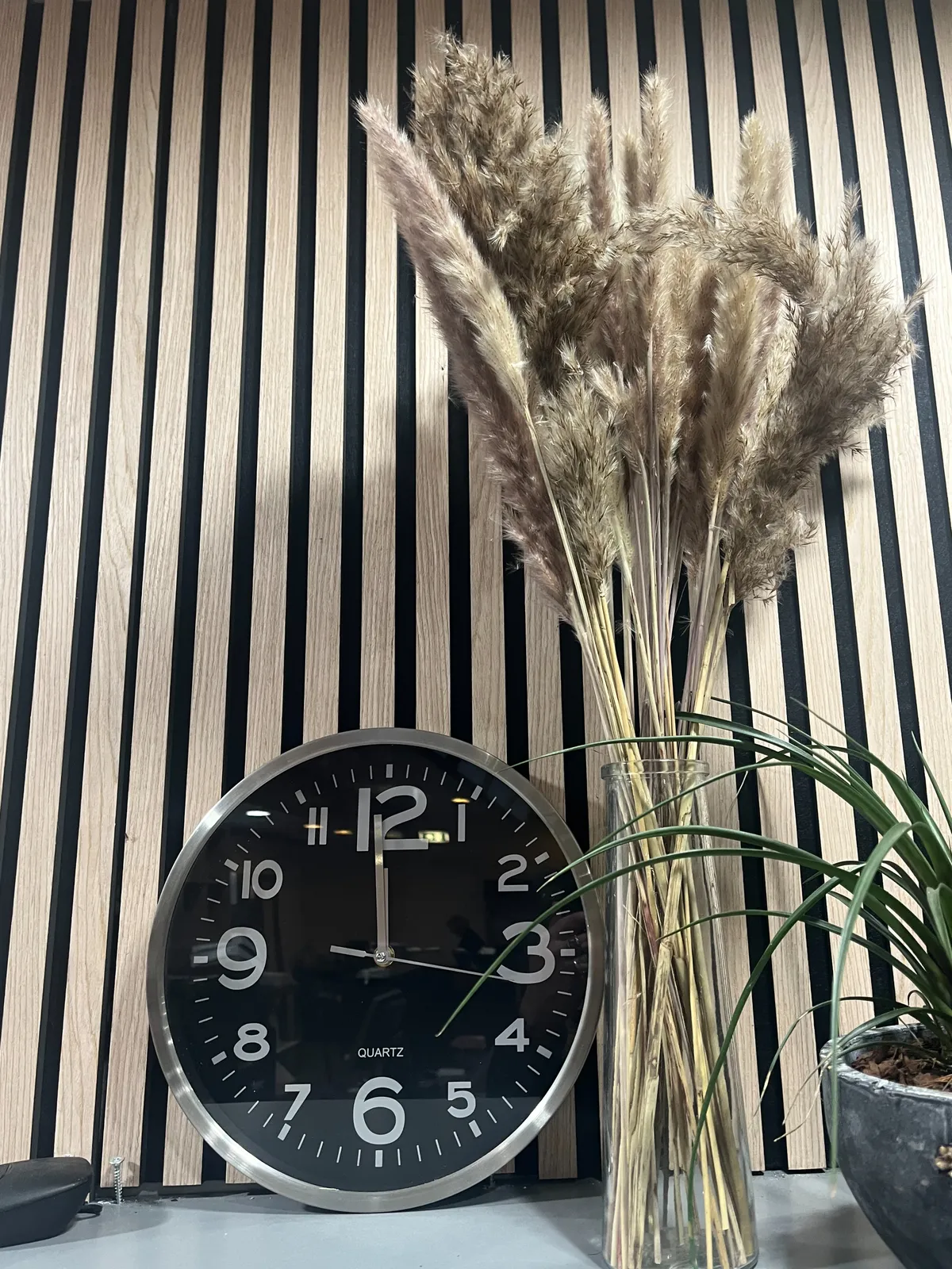In front of a vertically striped background, next to a vase with plumes, there’s a clock that points at one minute to 12.
