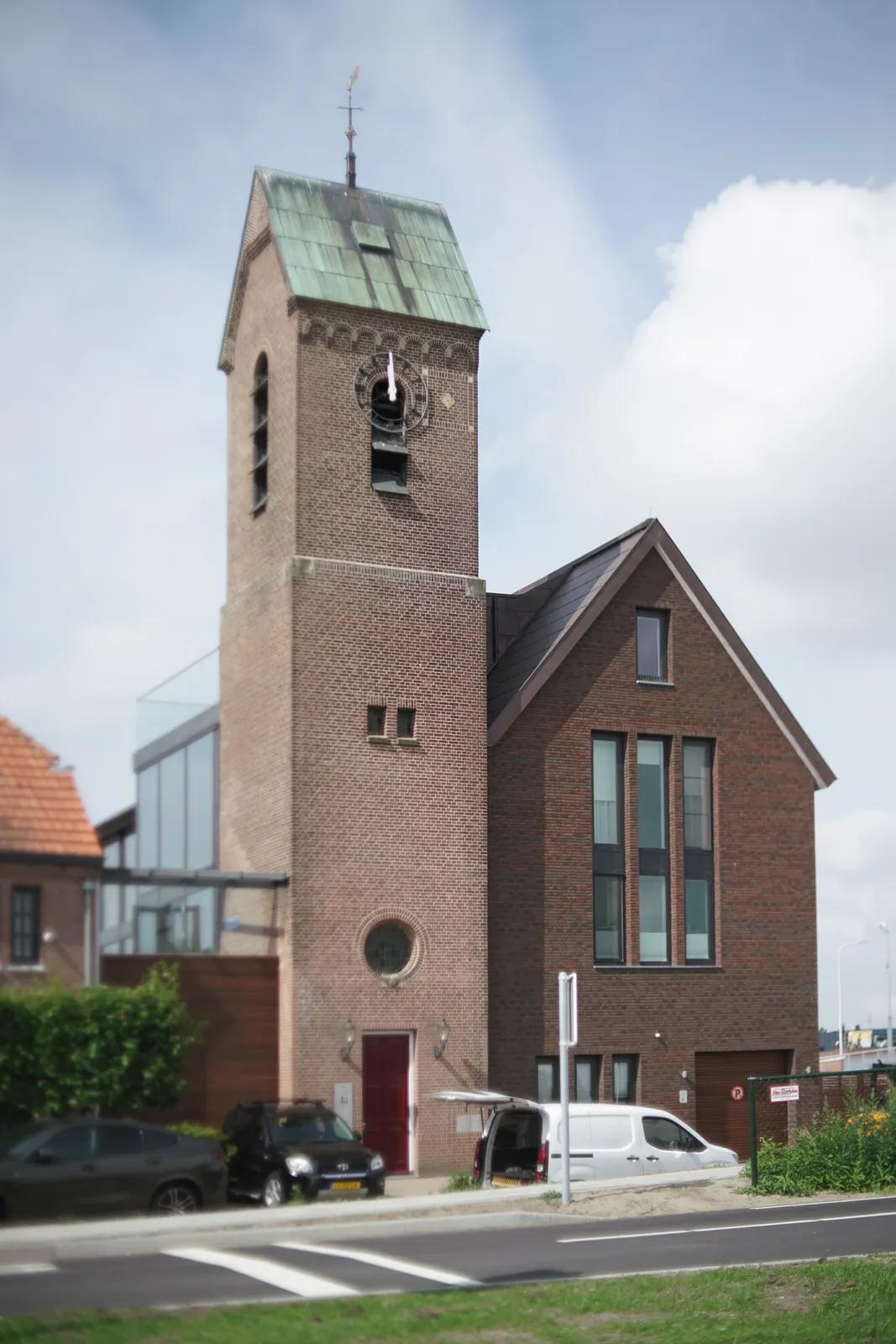 An eclectic combination of a seemingly old church tower and a modern building. On the church there’s a clock that points at 1 minute to 12.  