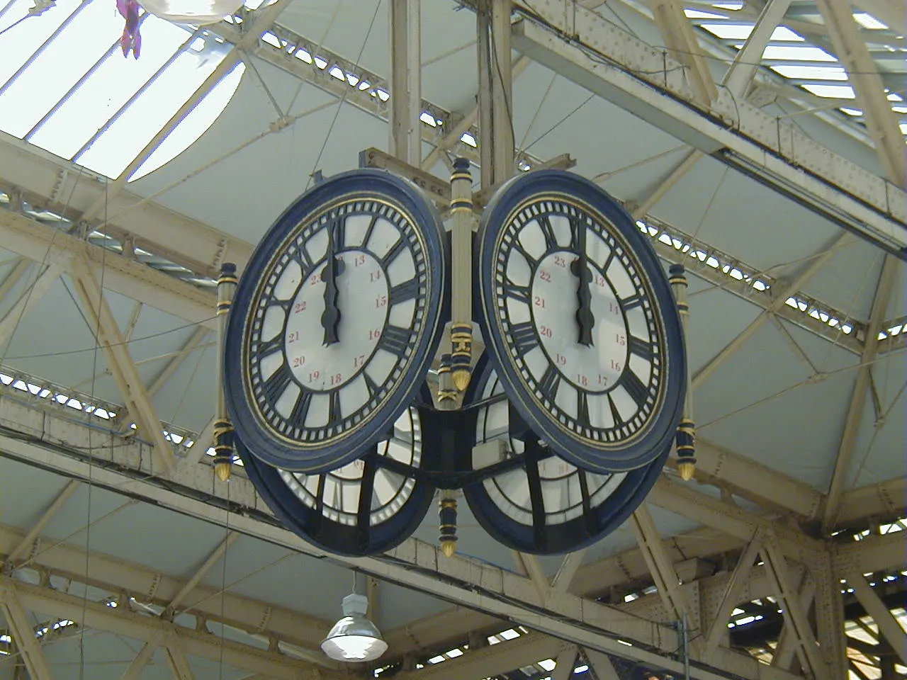 Four large clocks hanging from a ceiling in a huge hall. The two clocks that are visible point at noon.
