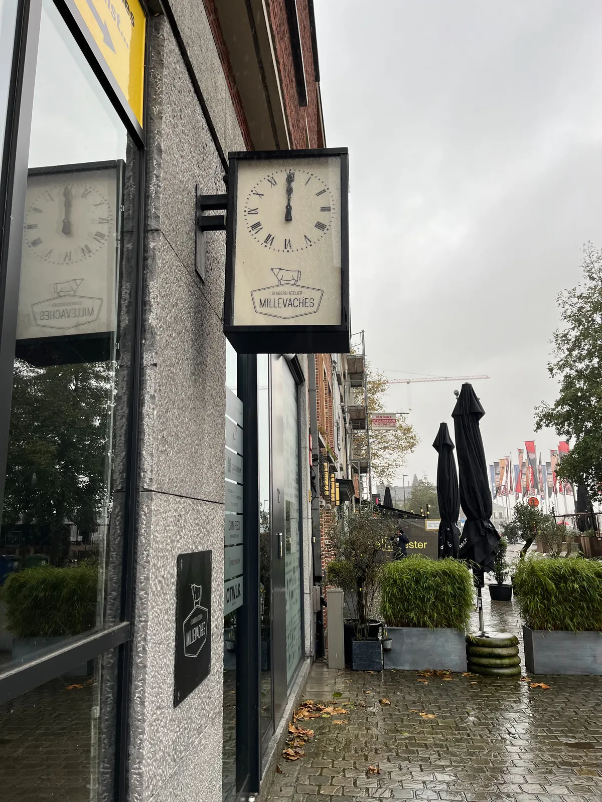 A clock attached to a shop in a rainy city. It points at exactly twelve o’clock.