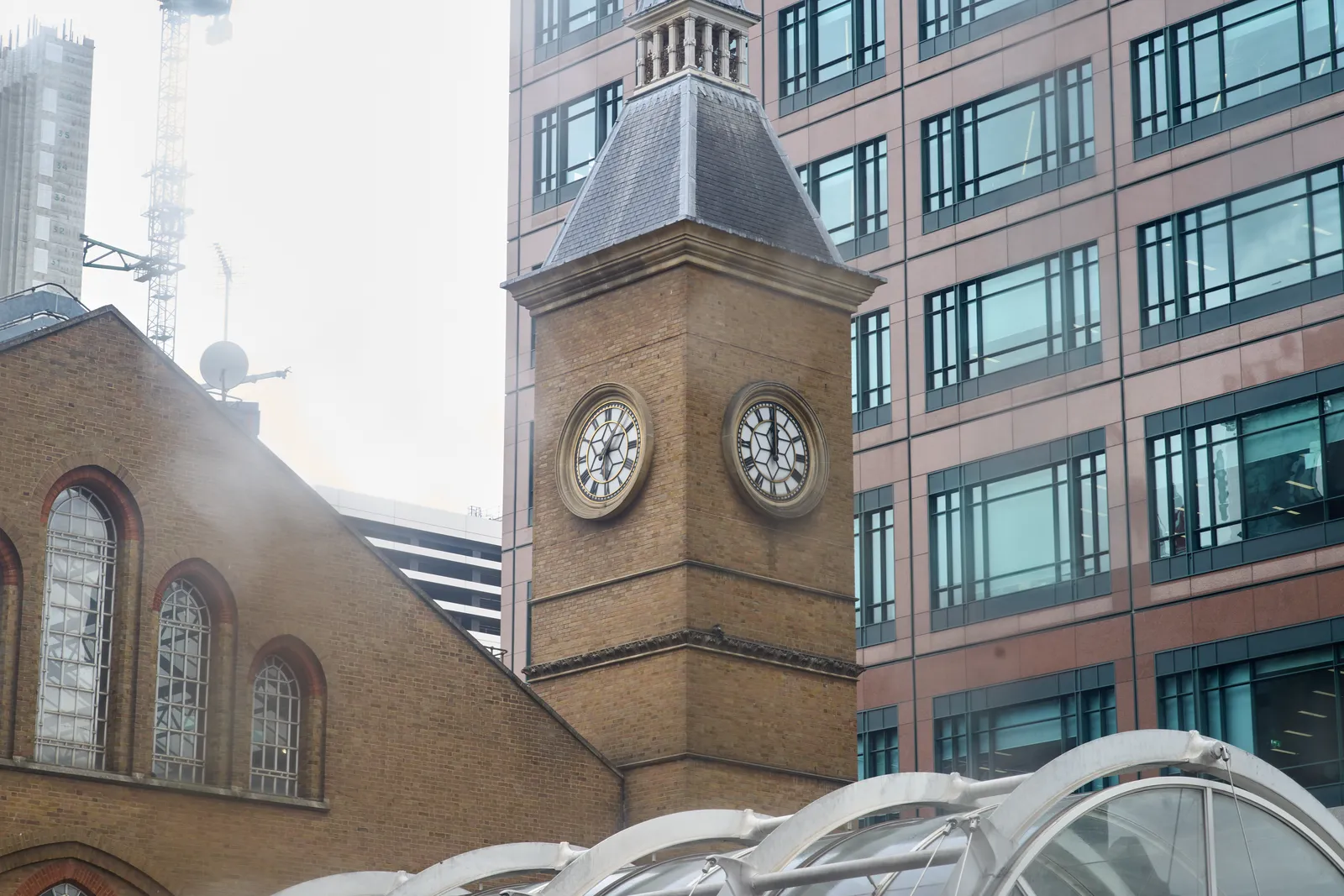 An older brick building with a tower with on two sides, a visible clock on it, surrounded by much higher, more modern buildings. One of the clocks points at 6 minutes past six, the other at twelve o’clock.