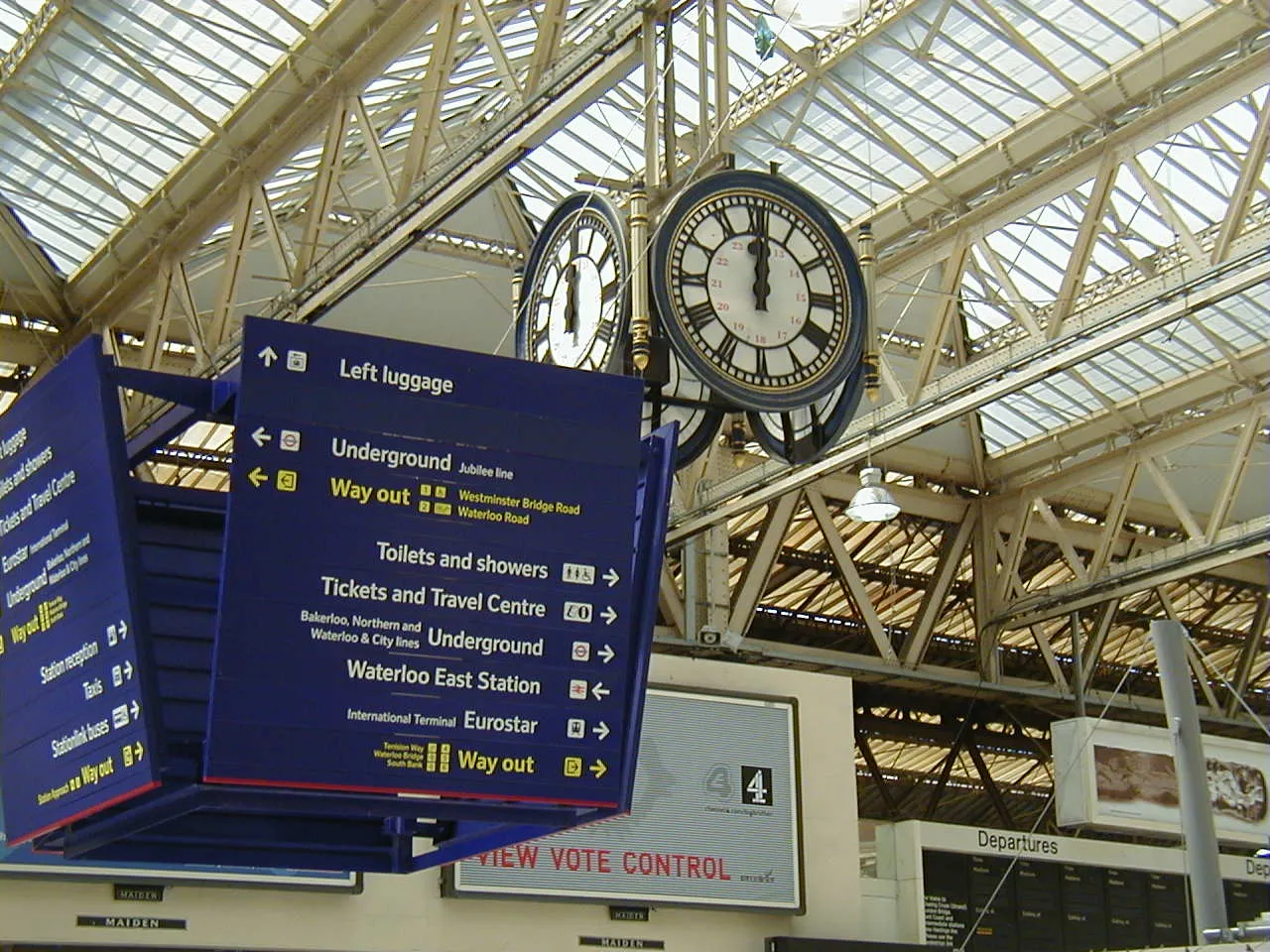 An enormous clock, hanging from a ceiling in a train station points at two minutes past 12.