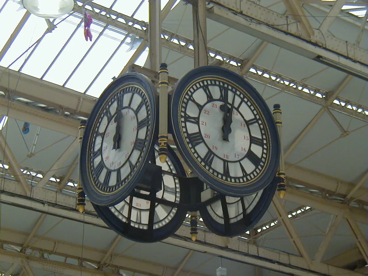 An enormous clock, hanging from a ceiling in a train station points at three minutes past 12.
