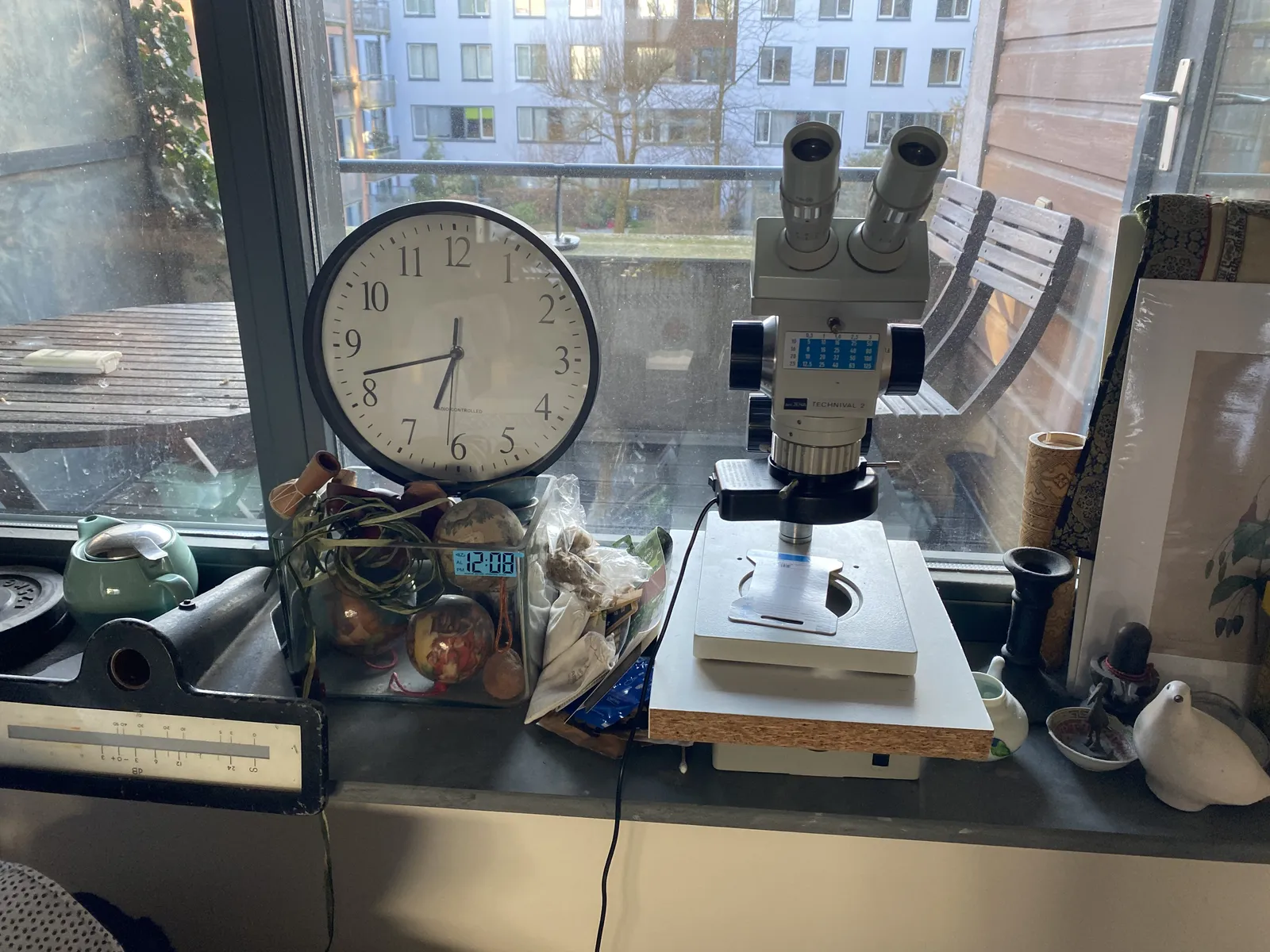 There’s a microscope on this image. Left of it is a glass vase with a sticker on it with the timestamp 12:08. Which it is right now. On top of the vase there’s a clock which points at 18 to seven, which it is not.
