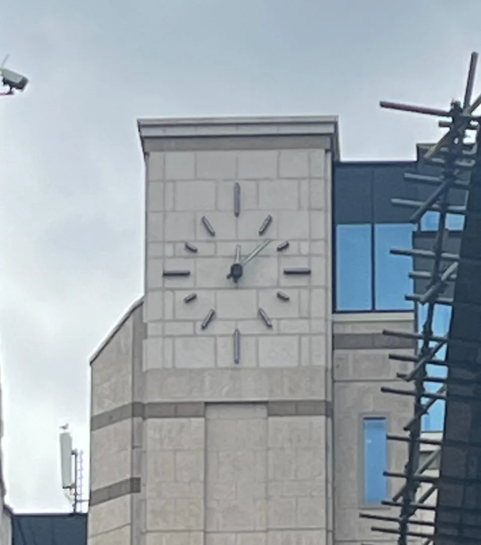 A modernist clock built into the facade of a modernist building. It points at 12 minutes past 12.