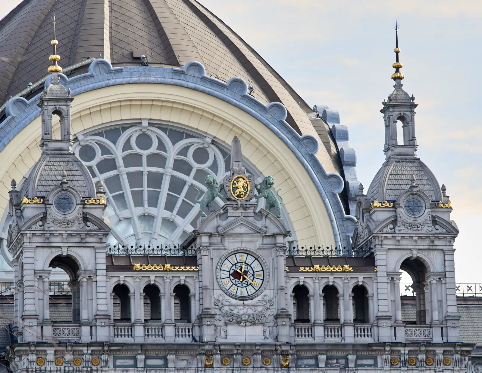 The dome of the Antwerp train station, with some decorated, and gold plated towers. On the center tower there’s a clock that points at 21 minutes past twelve