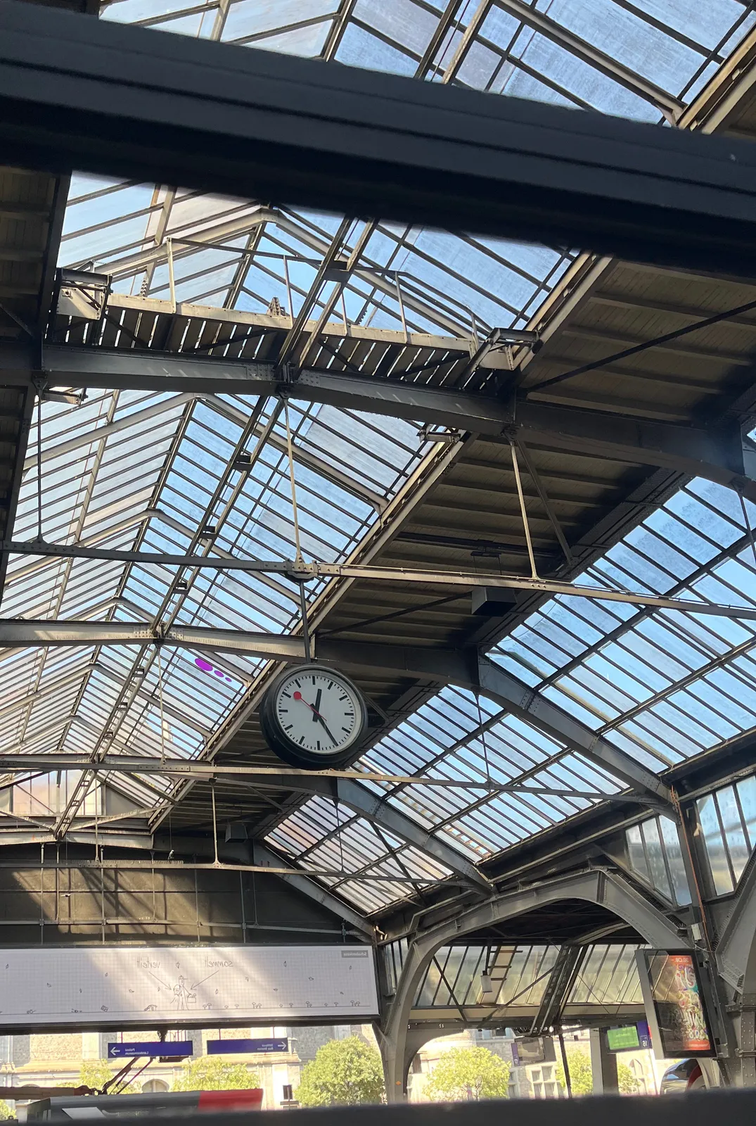 A clock on a station, between the large metal constructions of the roof. It points at 25 past 12.