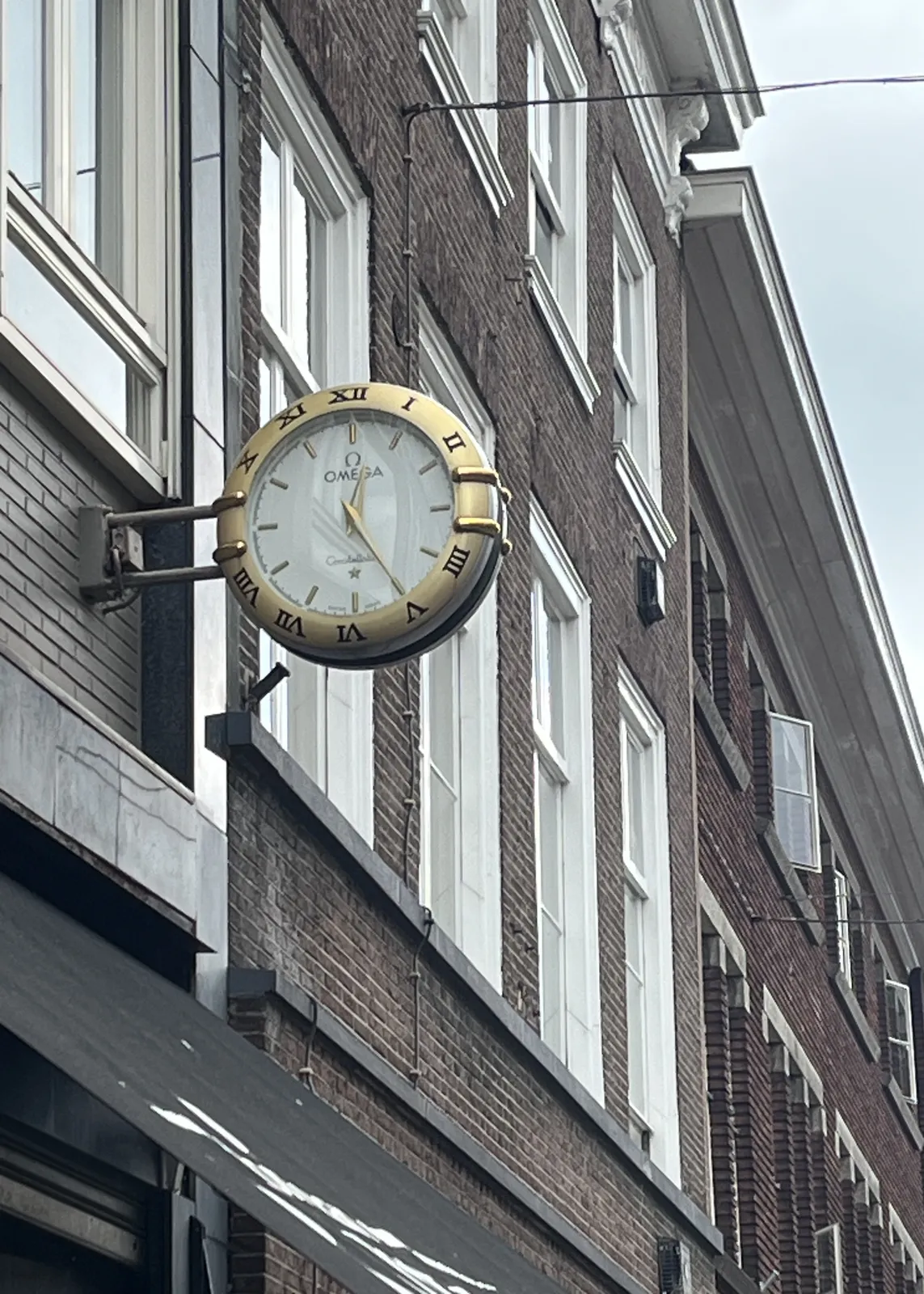A clock attached to a brick building. It is shaped like a posh omega wrist watch. It points at 25 past twelve.