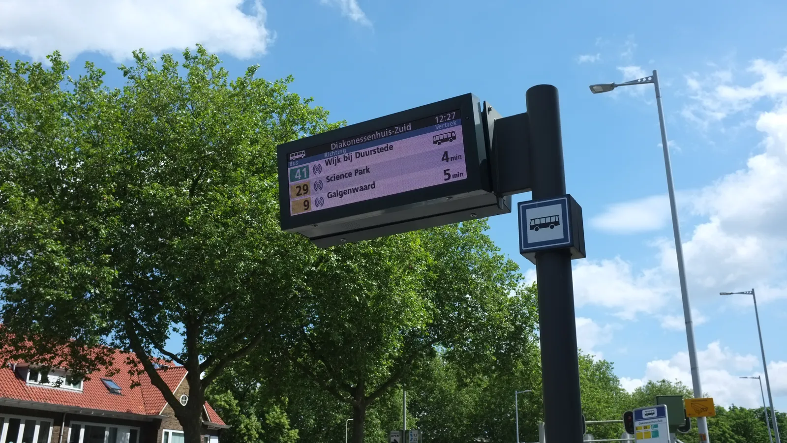 A digital display at a busstop. It shows a few busses that are about to arrive, and it shows the time as 12:27.