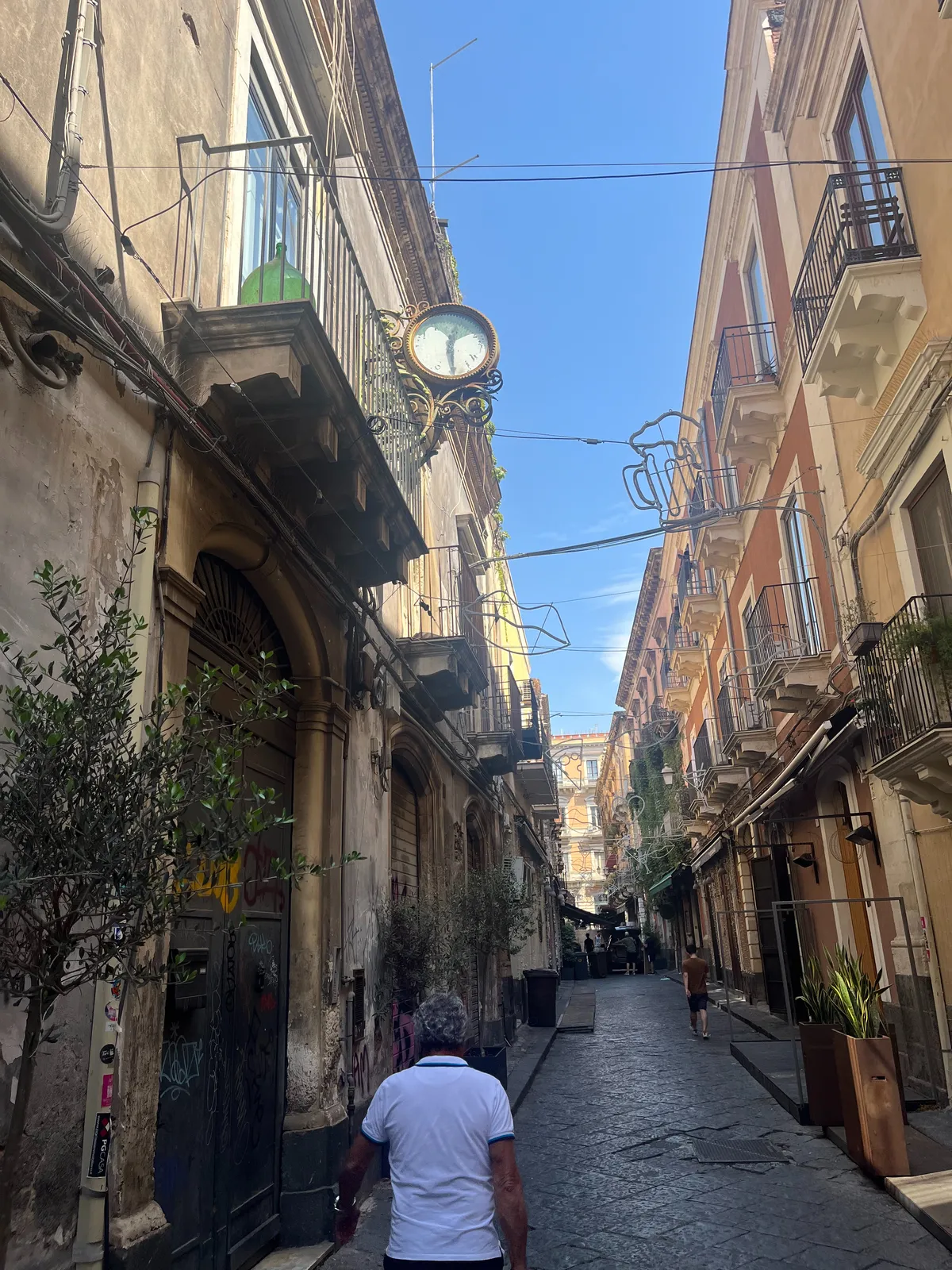 A wonderful summery road with nice curly balconies and a curly, very old clock attached to the facade of one of the houses. The clock points at half past twelve.