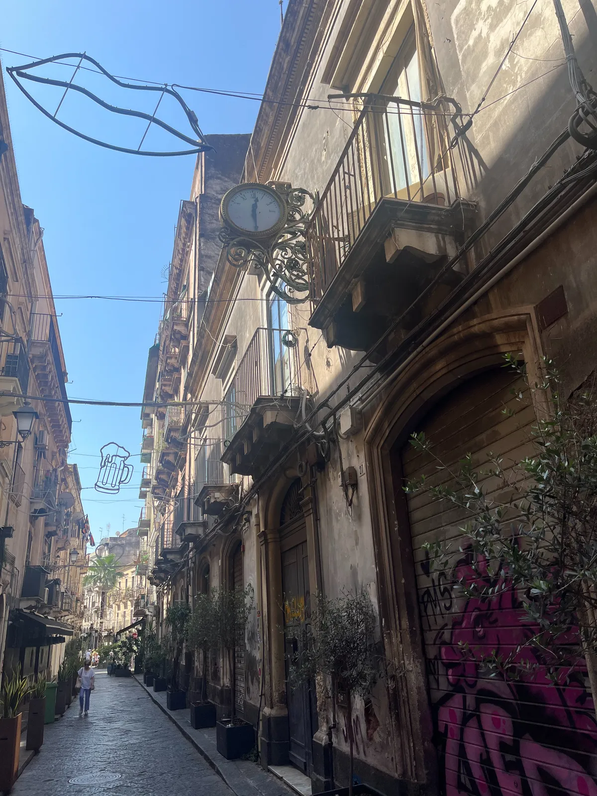 A wonderful summery road with nice curly balconies and a curly, very old clock attached to the facade of one of the houses. The clock points at half past twelve.