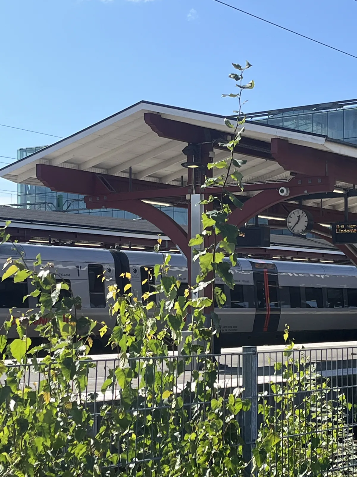 Looking past a fence, past some shrubbery at a train, parked on a platform. The clock on this platform points at 37 minutes past 12.