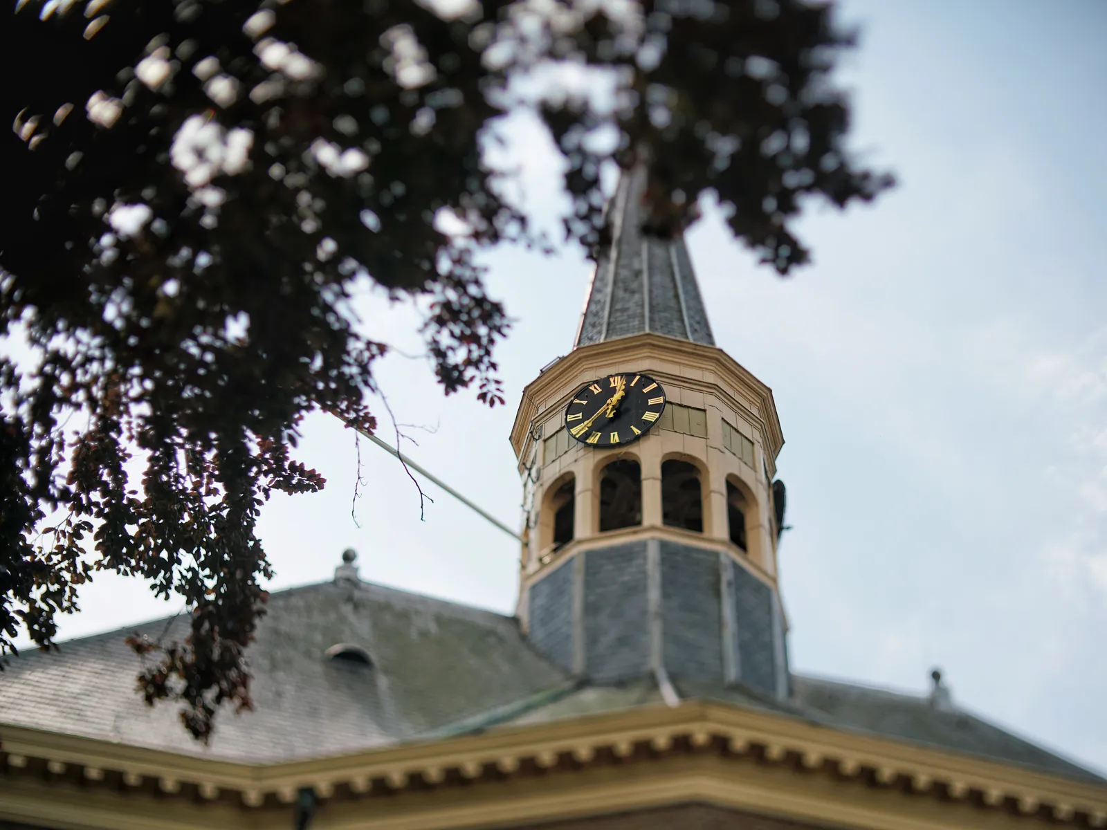 Peeking through some trees at an old church roof with a little tower on it. On the tower there’s a clock that points at 21 minutes to one.