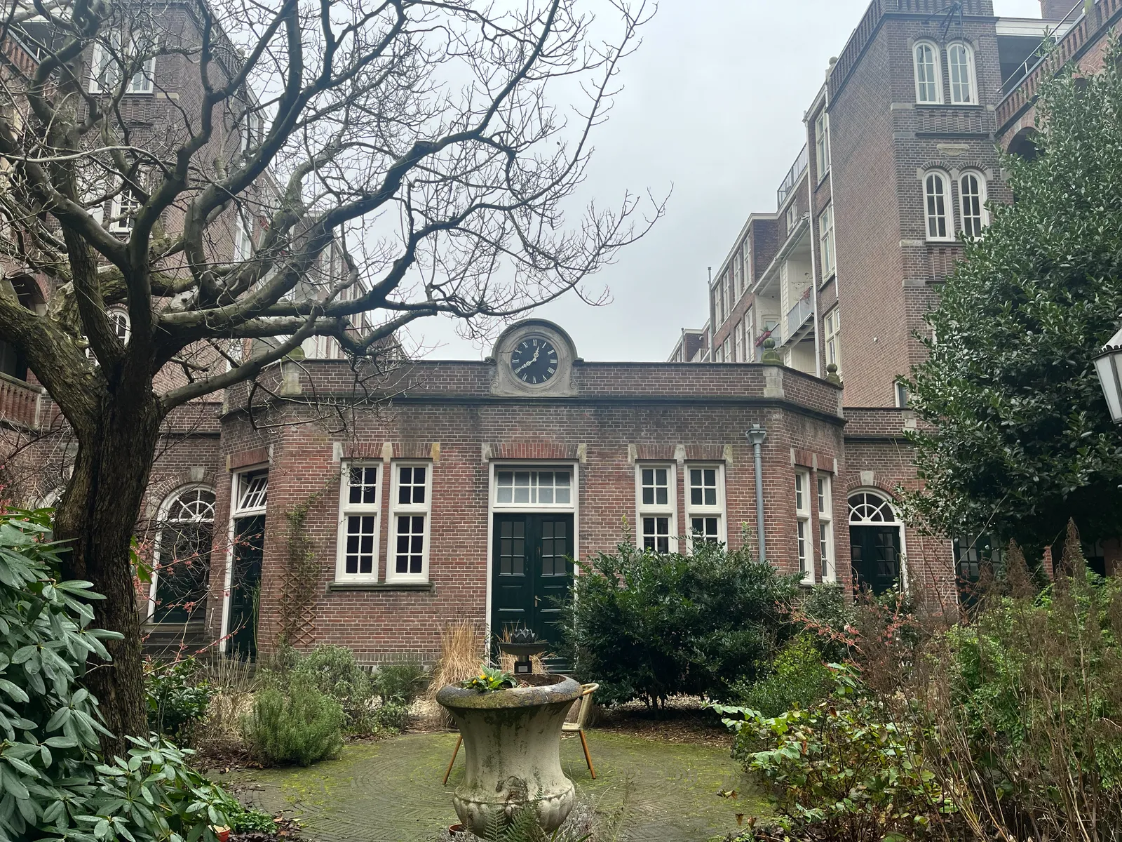 A garden with a tree and some other plants. It seems like it’s winter, there are no leaves on the tree. There’s also a brick building in this garden with a clock on it. It points at 20 minutes to one.