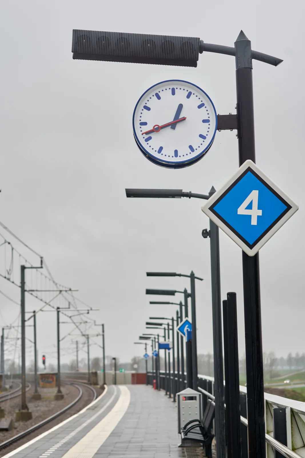 On a train station. The track can be seen, and quite a few street lights on the platform. Attached to one of these lights there a clock that points at 42 past 12.