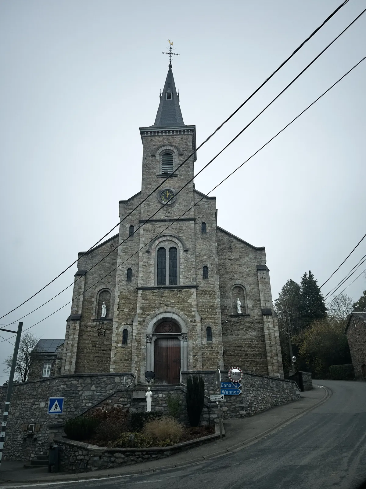 Looking up at an old stone church. According to the signs, the road to Ennal and Wanne goes past it. The clock on the church points at one o’clock.