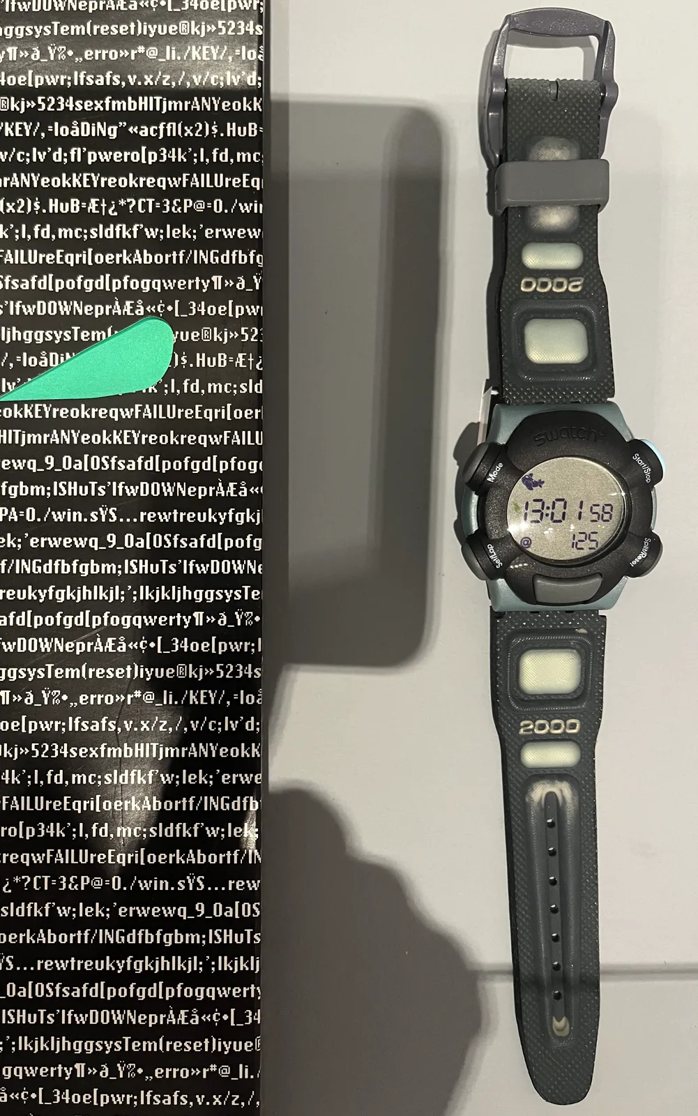 A bulbous, black and gray wrist watch with the number 2000 printed on the strap. It has a digital display which shows the time in two different ways. It shows it in the usual 24 hour time, as 13:01, and it shows it in so called beat time. In beat time it is 125.