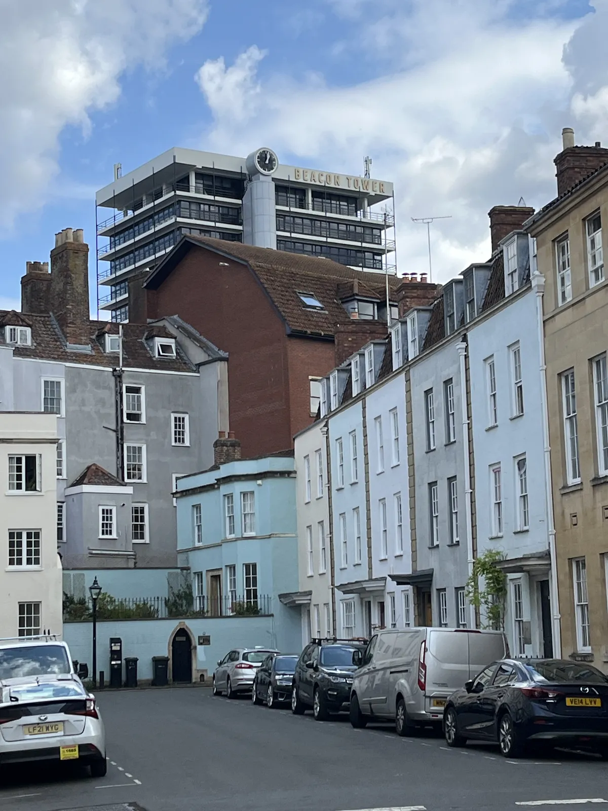 Parked cars in a dead end street with two, three and four storey buildings around it. Behind these buildings towers a very high flat, with a clock on top, which points at 3 minutes past three.