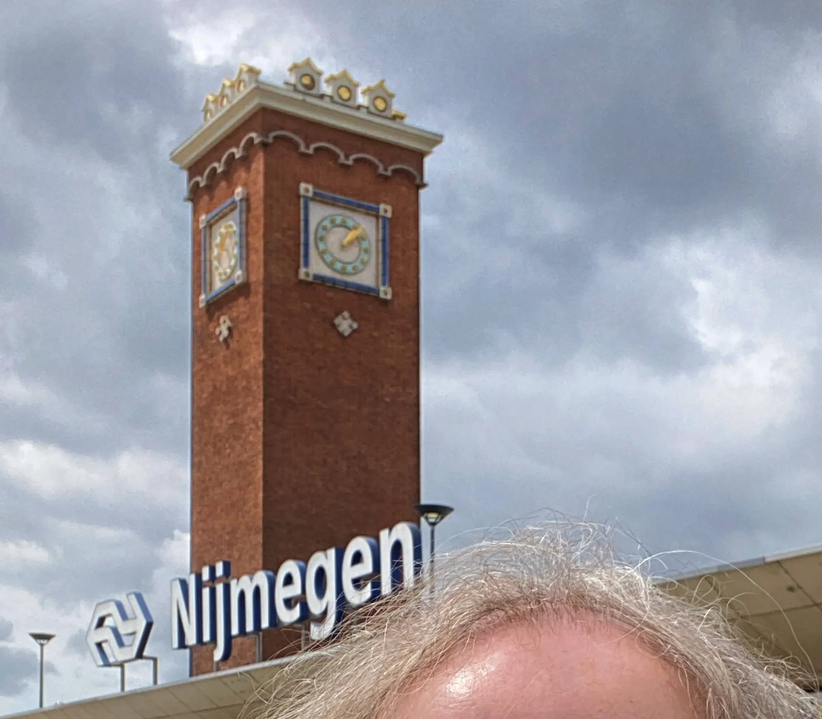 The clock on the tower of the station in Nijmegen points at approximately 7 past 1. In the foreground there’s a shiny head of a balding man.