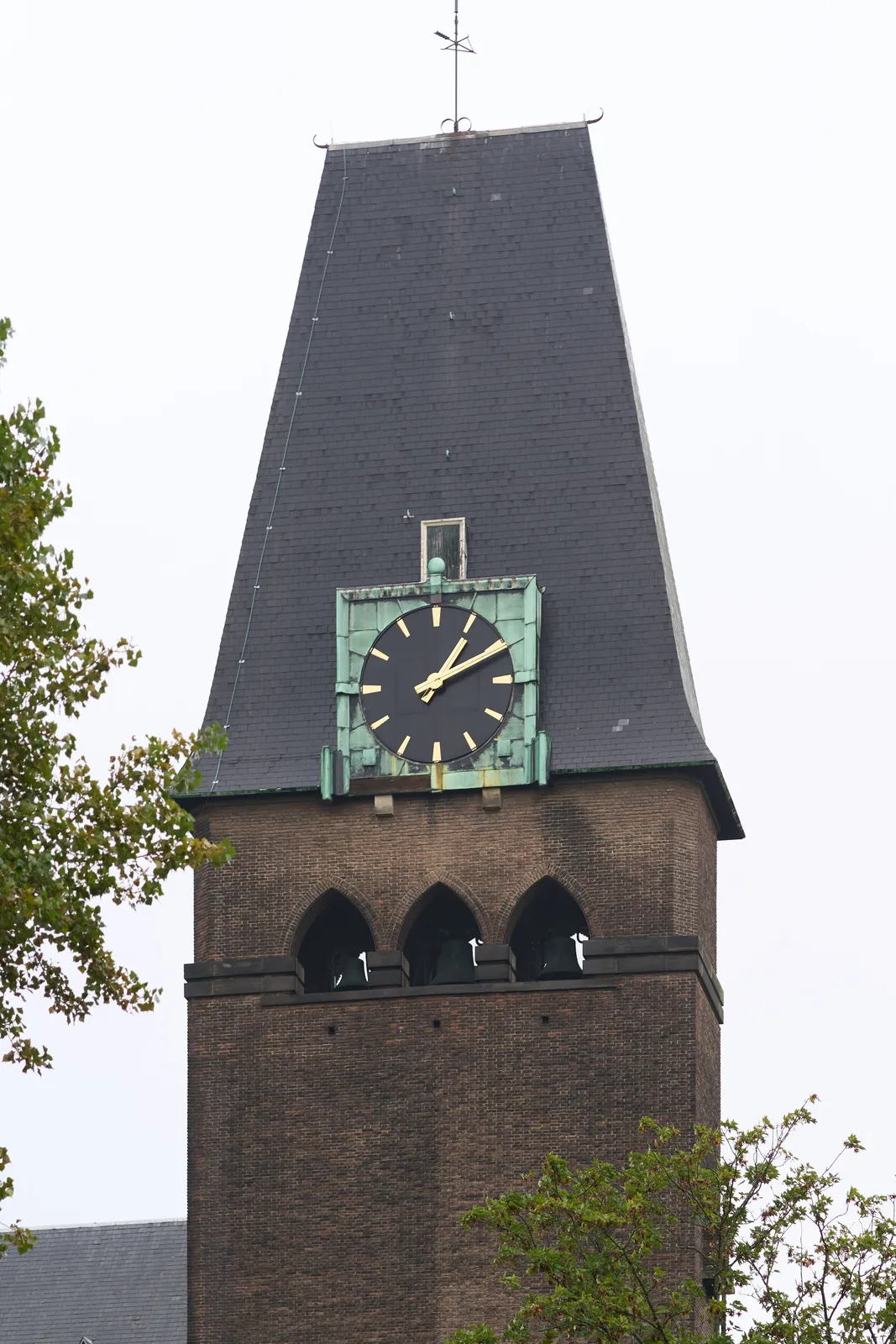 A monumental, slate-roofed, brick church tower, with a large brass clock on it. It points at 10 past one.