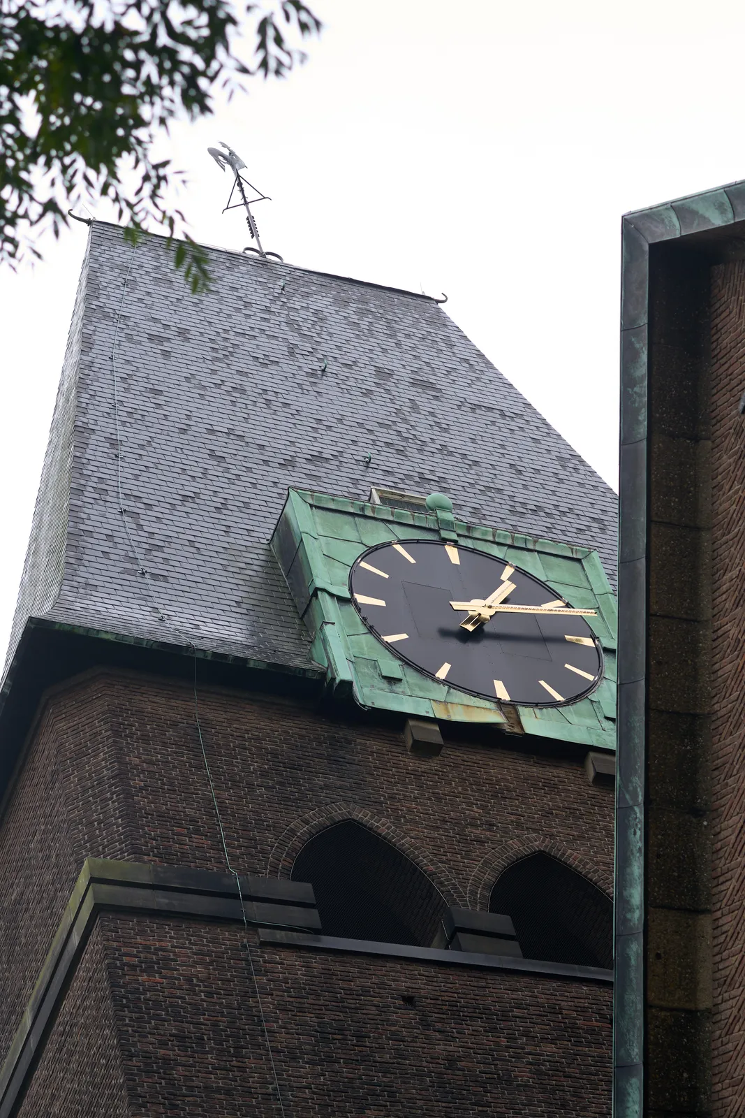 A picture taken from below, looking up at a monumental, slate-roofed, brick church tower, with a large brass clock on it. It points at 13 minutes past one.