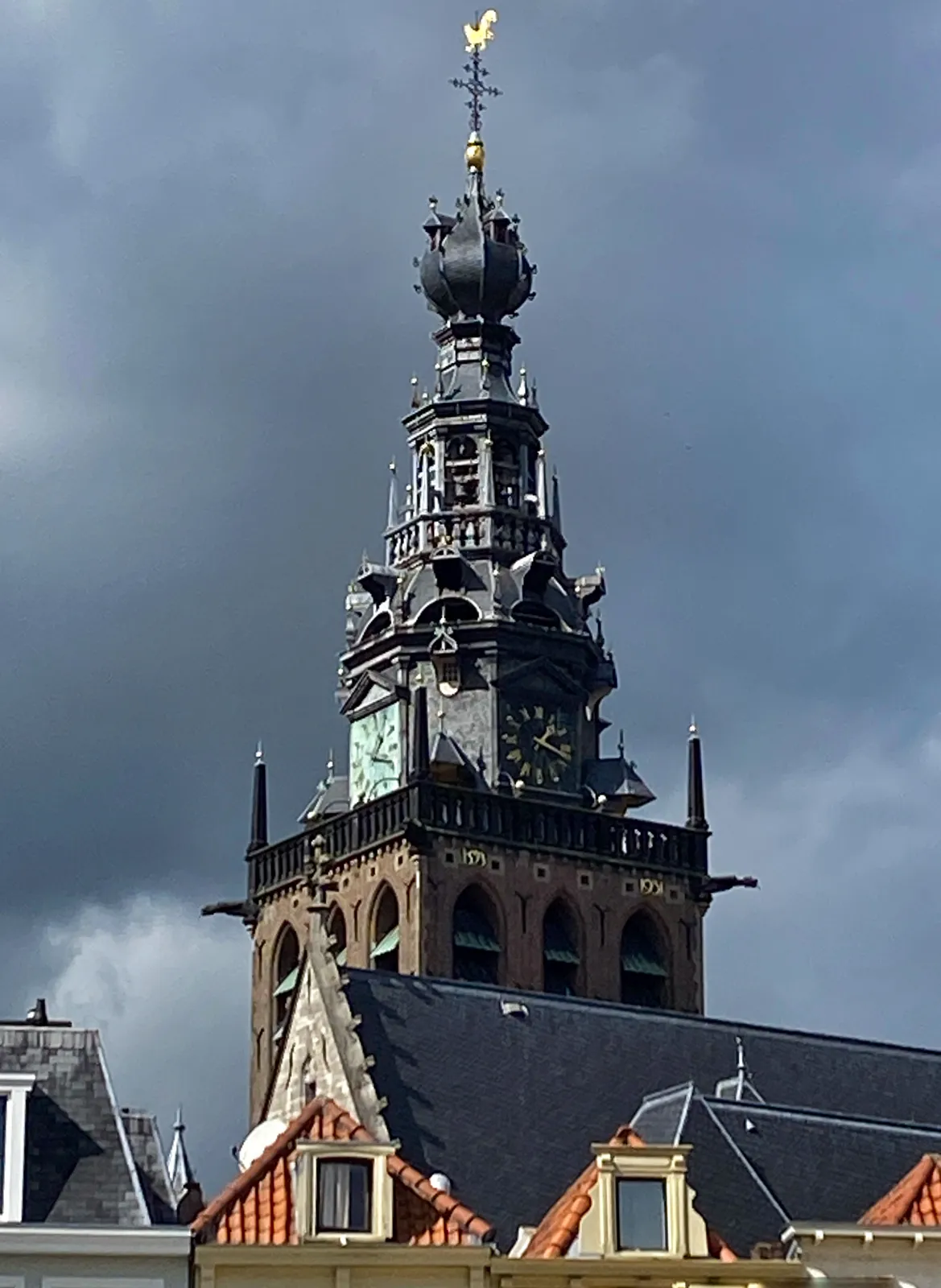 A richly ornamented old church tower, towering over some roofs. The tower is brightly lit by the sun, but it is set against a gray, rainy sky. The clock on the tower points at 17 minutes past one.