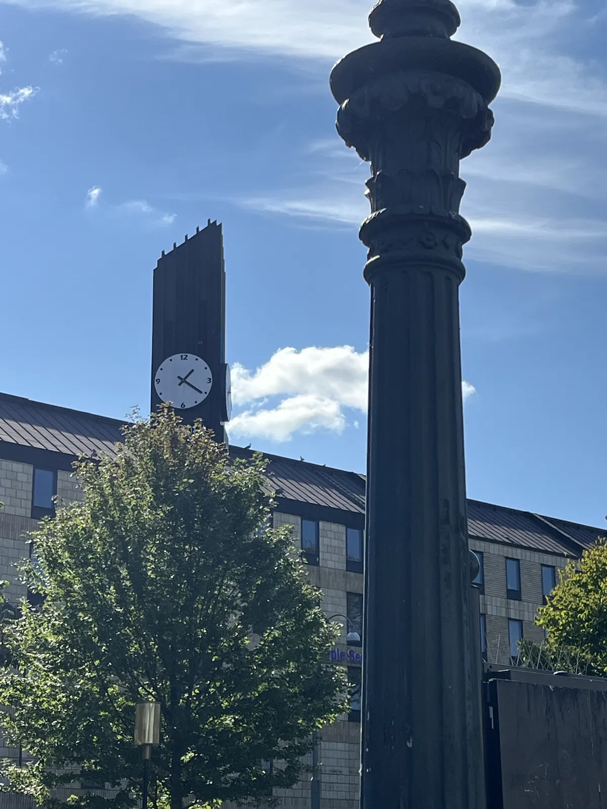 Looking past a lamp post, at a brick building, with a large tree in front of it. On top of the building there’s a modern tower with a clock on it that points at 20 past one