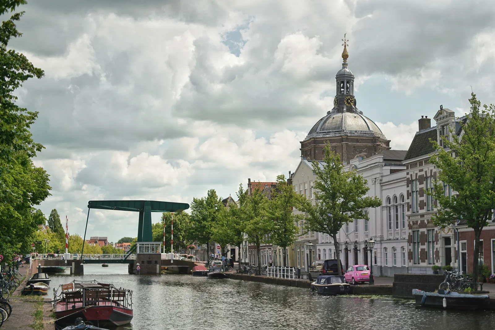 A canal view in Leiden, with houses, a bridge and a few boats and a pink car. Behind the houses there’s the dome of a large church, with a clock on top. The clock points at 23 minutes past one. 