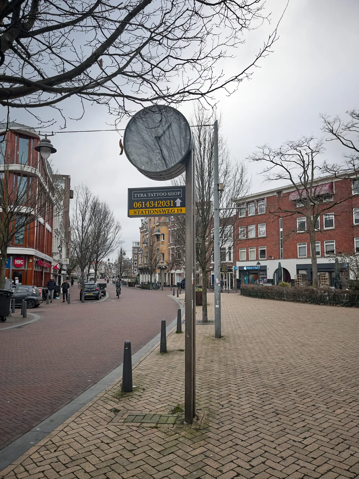 A blick street, with brick buildings. There’s a pole, with an advertisement for a tattoo shop, and a clock. The clock points at 27 minutes past 1.
