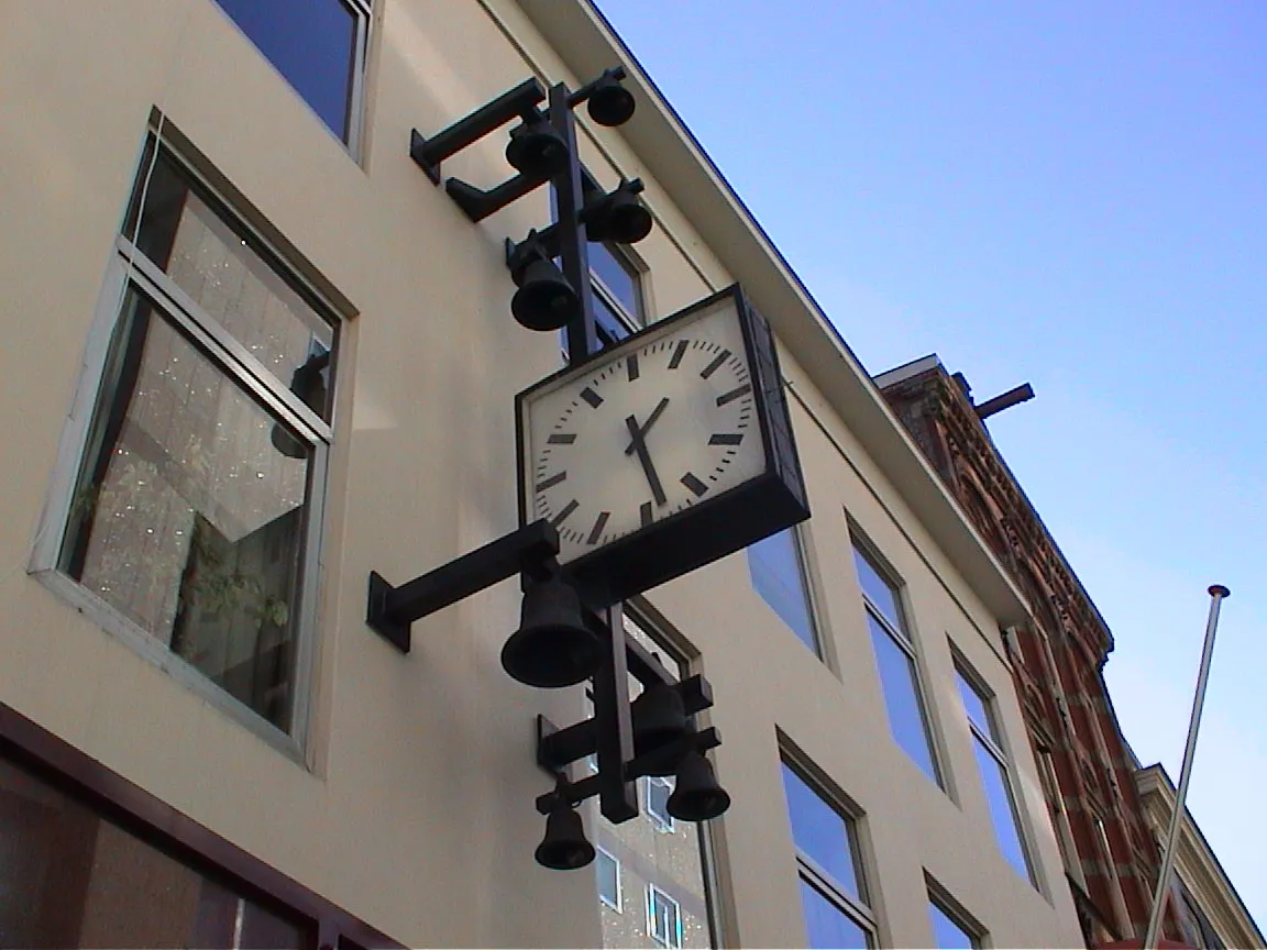 A modernist clock on a facade, surrounded by classic bells, points at 13:28